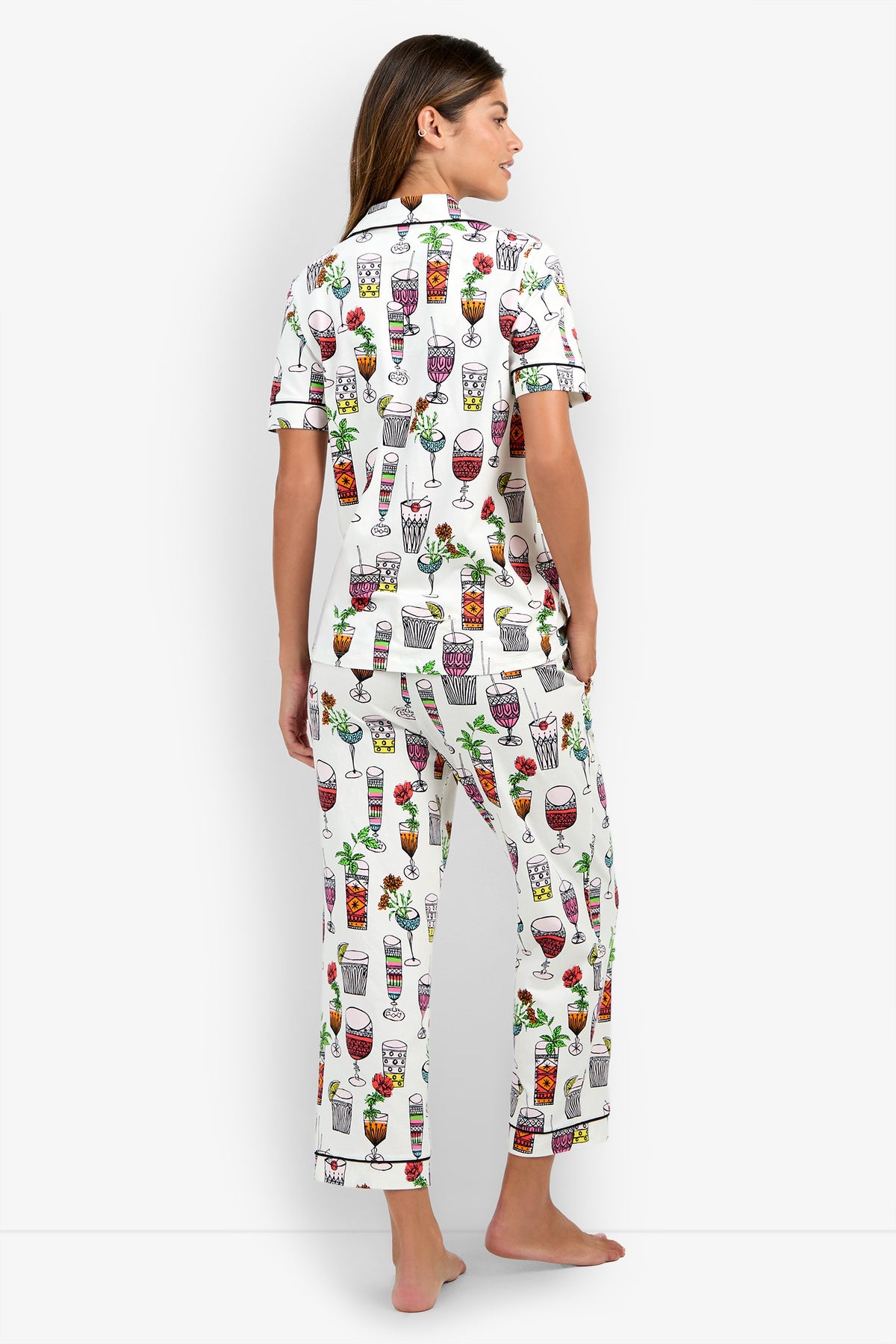 A woman stands barefoot, facing away, wearing the Short Sleeve Cropped Pajamas Set—white pajamas featuring colorful drink illustrations such as wine, cocktails, and juice glasses. The set includes a short-sleeve button-down top and long pants. The background is plain white.
