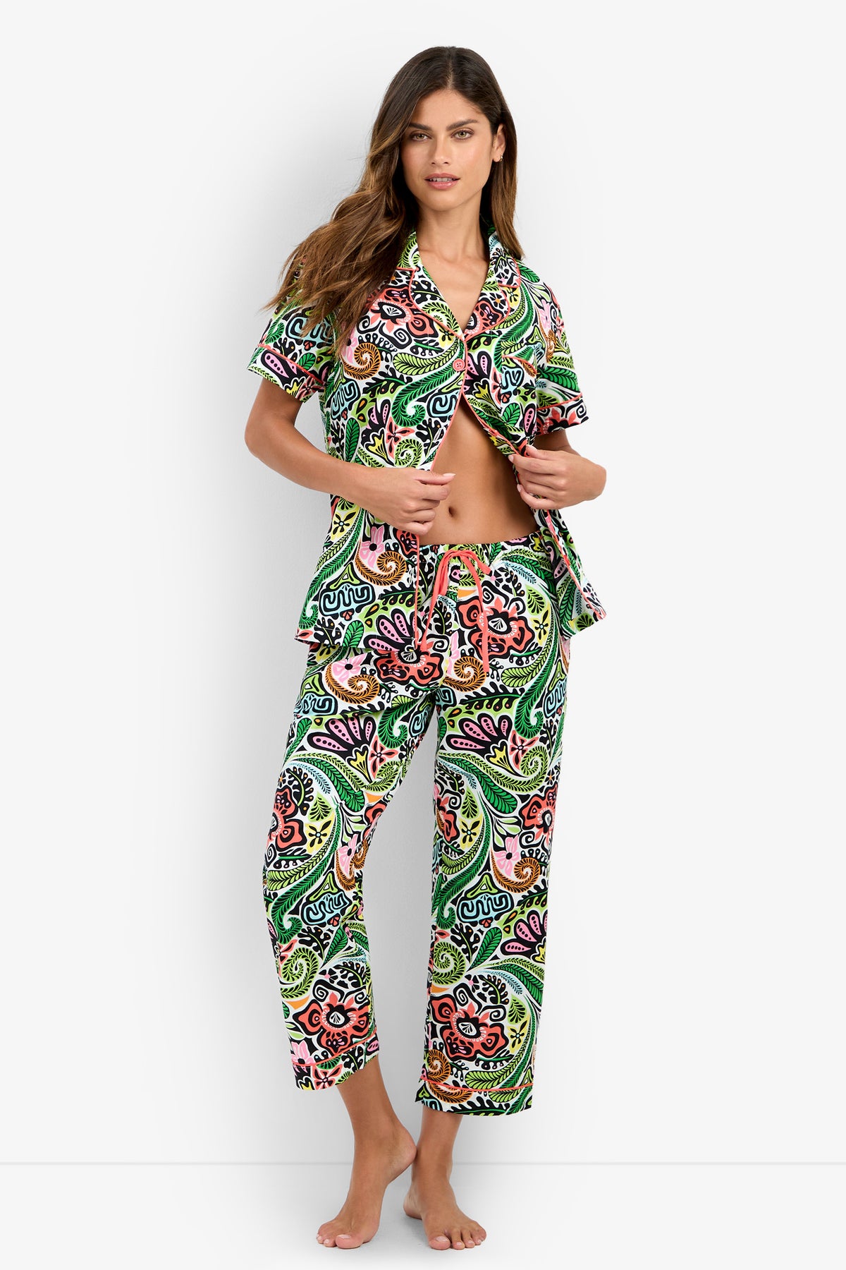 A woman stands barefoot against a plain white background, wearing the Short Sleeve Cropped Pajamas Set featuring a colorful paisley pattern, holding the shirt open to reveal her midriff.