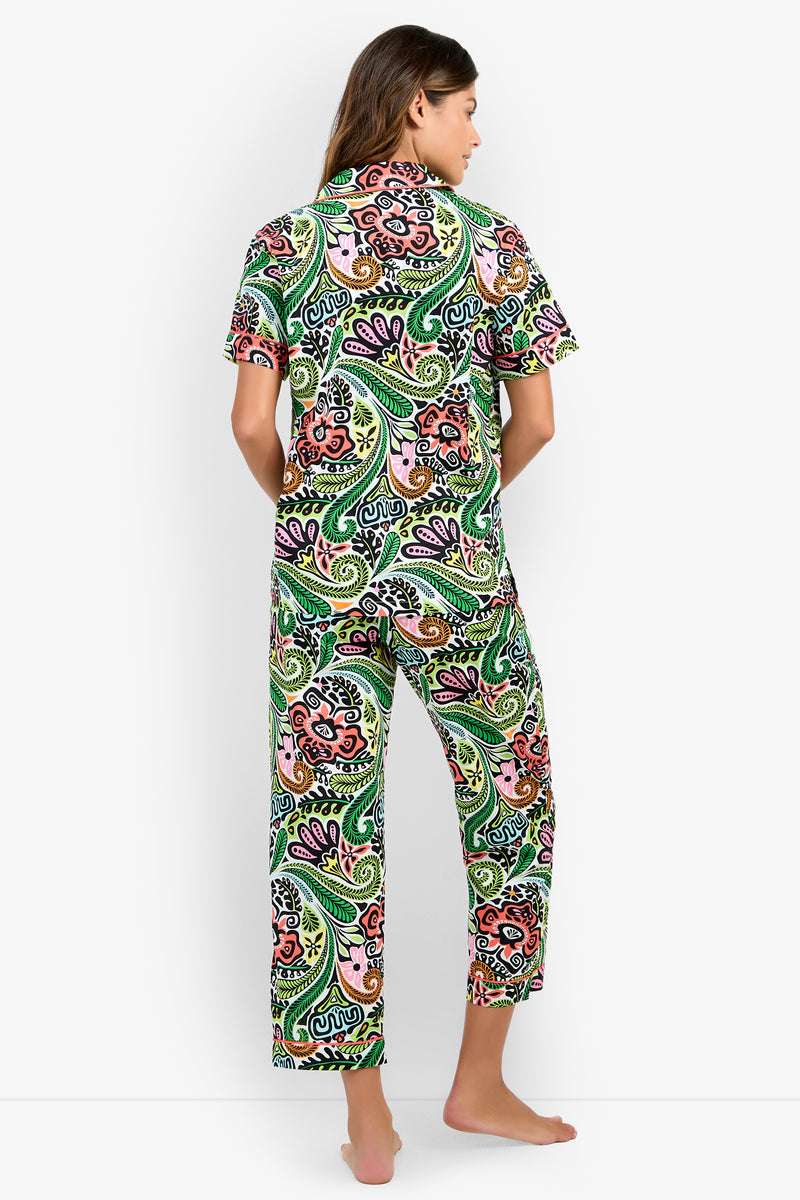 A woman stands barefoot, facing away, wearing the Short Sleeve Cropped Pajamas Set featuring a colorful paisley print with short sleeves and long pants. The background is plain white.