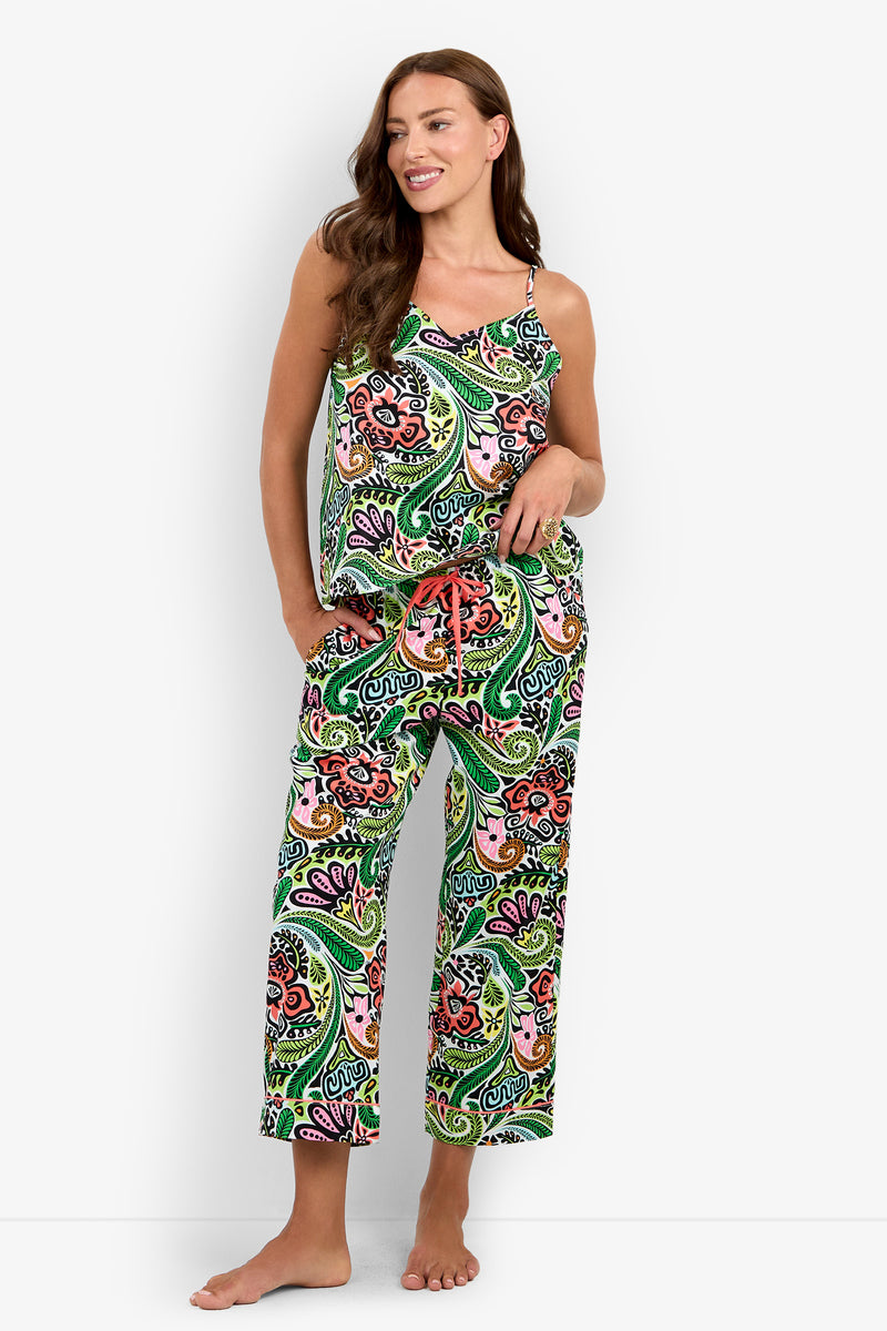 A woman stands barefoot and smiling against a plain white background, wearing the Tiki Cami Cropped PJ Set—a colorful, paisley-patterned pajama set featuring a sleeveless top and loose pants.