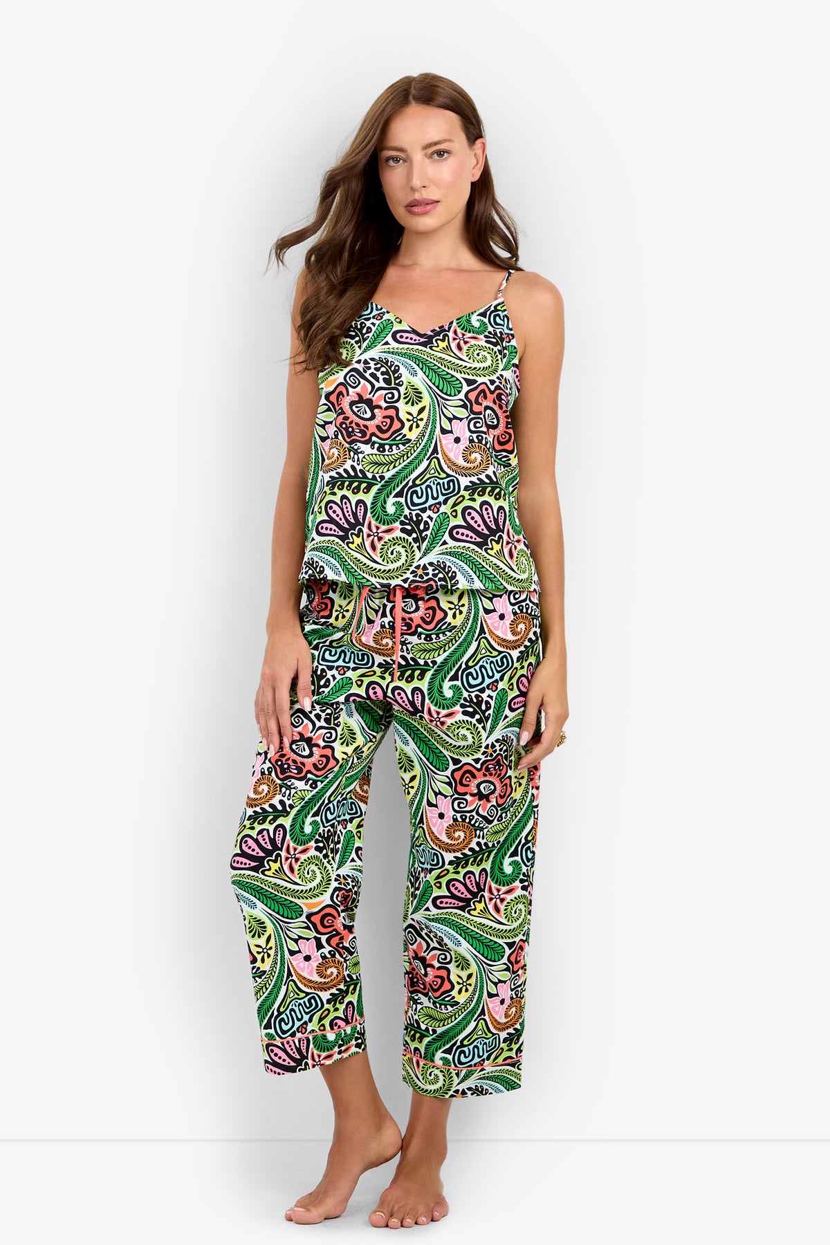 A woman wearing the Tiki Cami Cropped PJ Set, featuring a sleeveless top and wide-leg pants with a vibrant green, orange, purple, and black paisley pattern, stands barefoot against a plain white background.