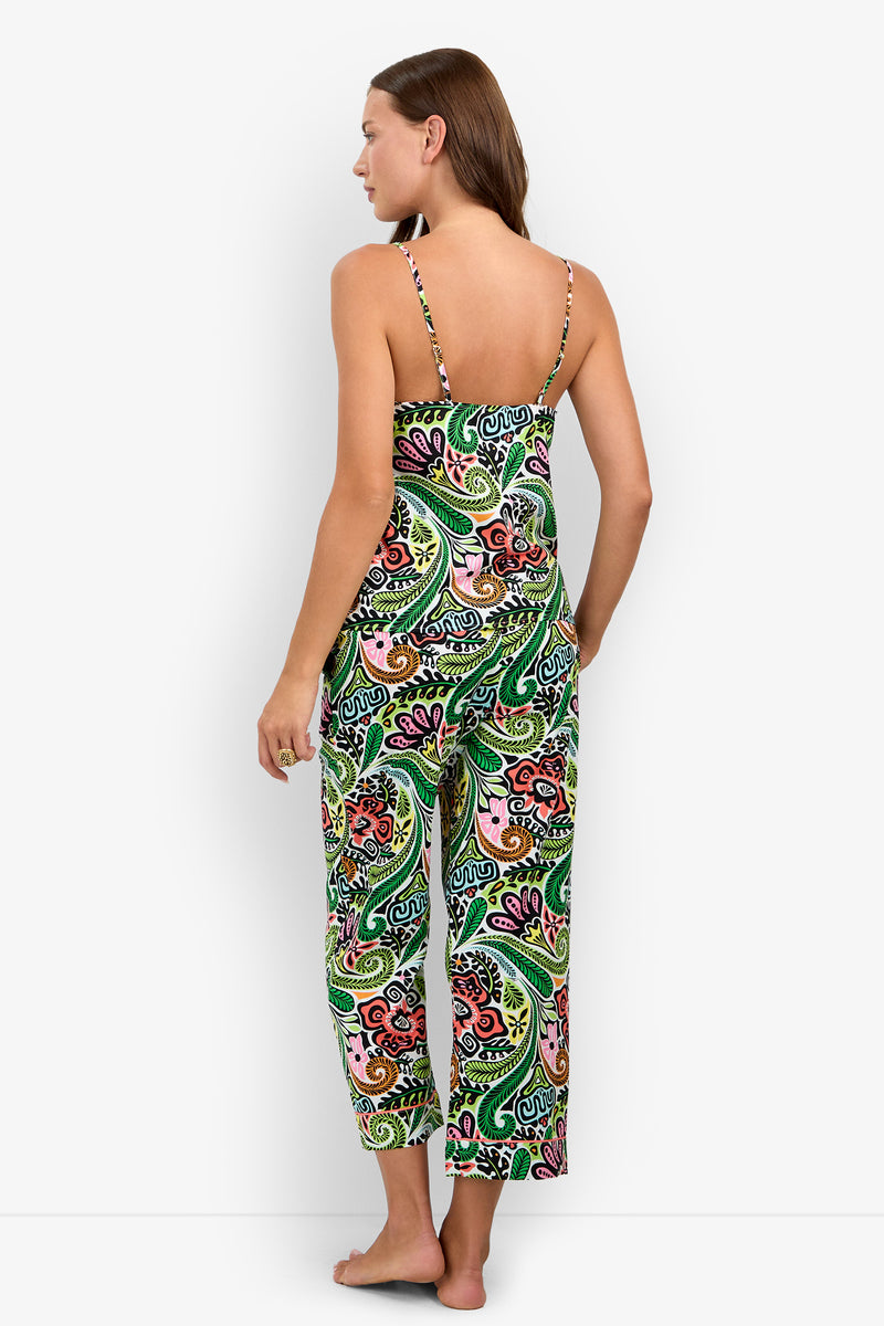 A woman stands barefoot facing away, wearing the Tiki Cami Cropped PJ Set—a colorful, paisley-patterned sleeveless jumpsuit with spaghetti straps. The background is plain white.