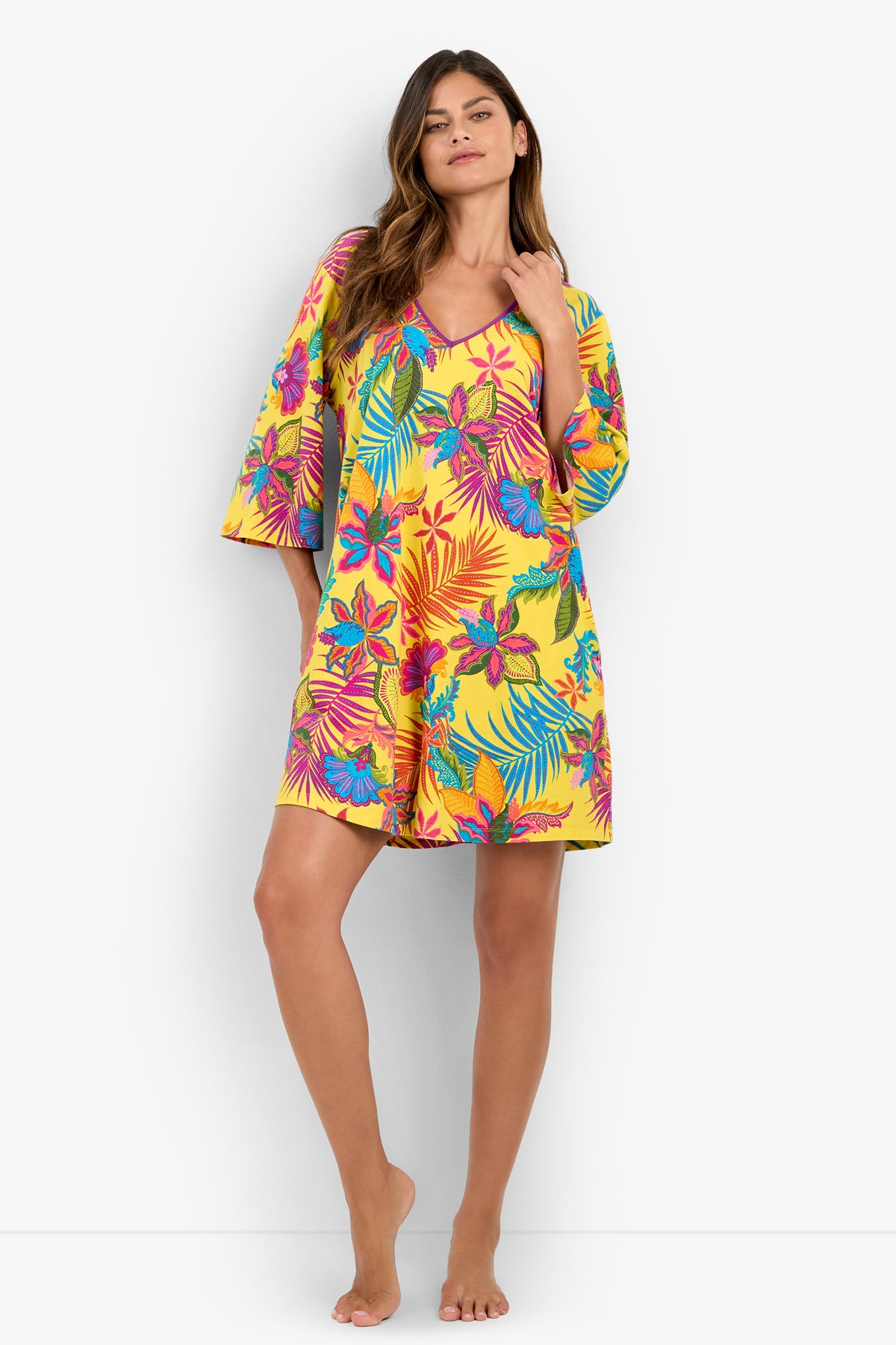 A woman stands barefoot wearing a loose, yellow Sleepshirt adorned with bright blue, pink, and green tropical floral patterns. She has long hair, touches her neck with one hand, and gazes confidently at the camera.