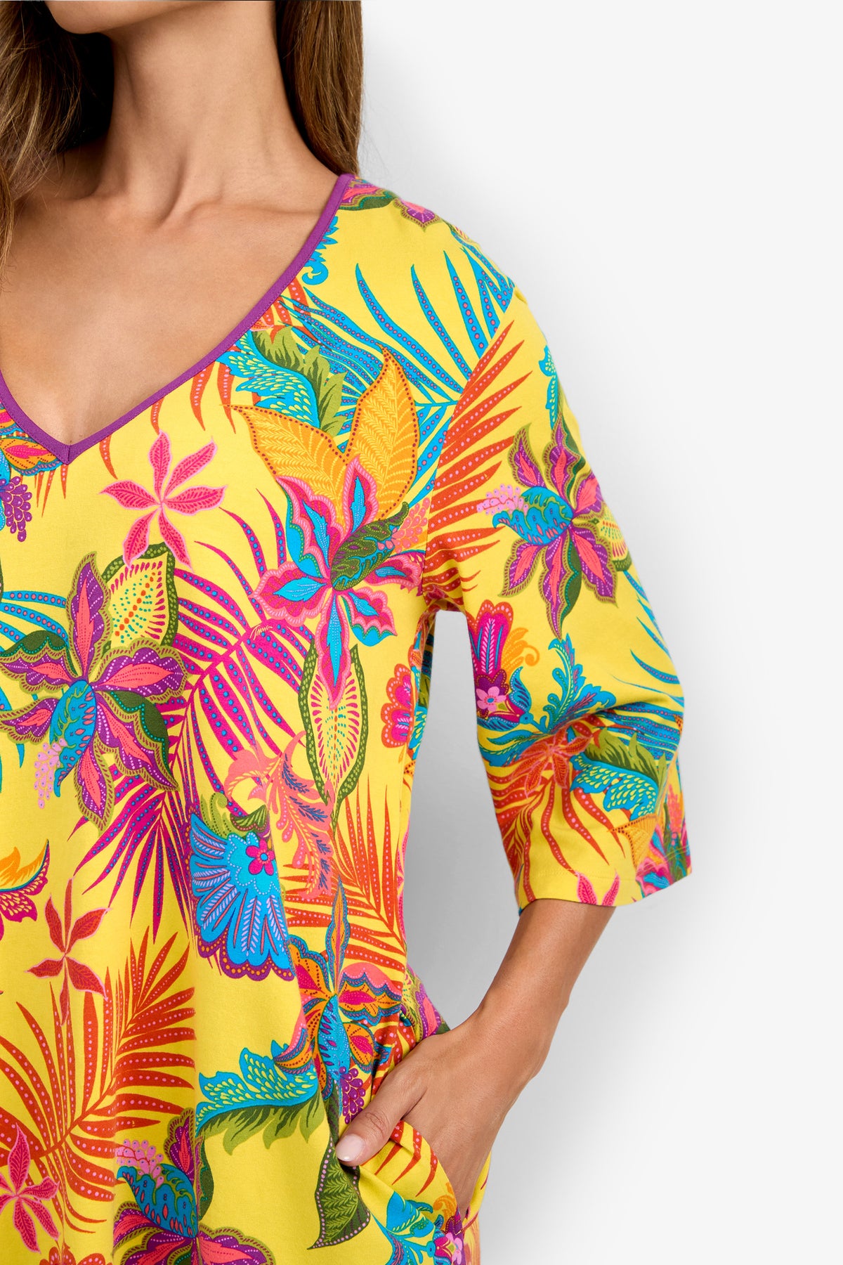 A woman wearing a vibrant yellow sleepshirt with colorful tropical floral patterns featuring pink, blue, and green leaves. The sleepshirt has a V-neck and elbow-length sleeves, and the woman’s hand is in her pocket.