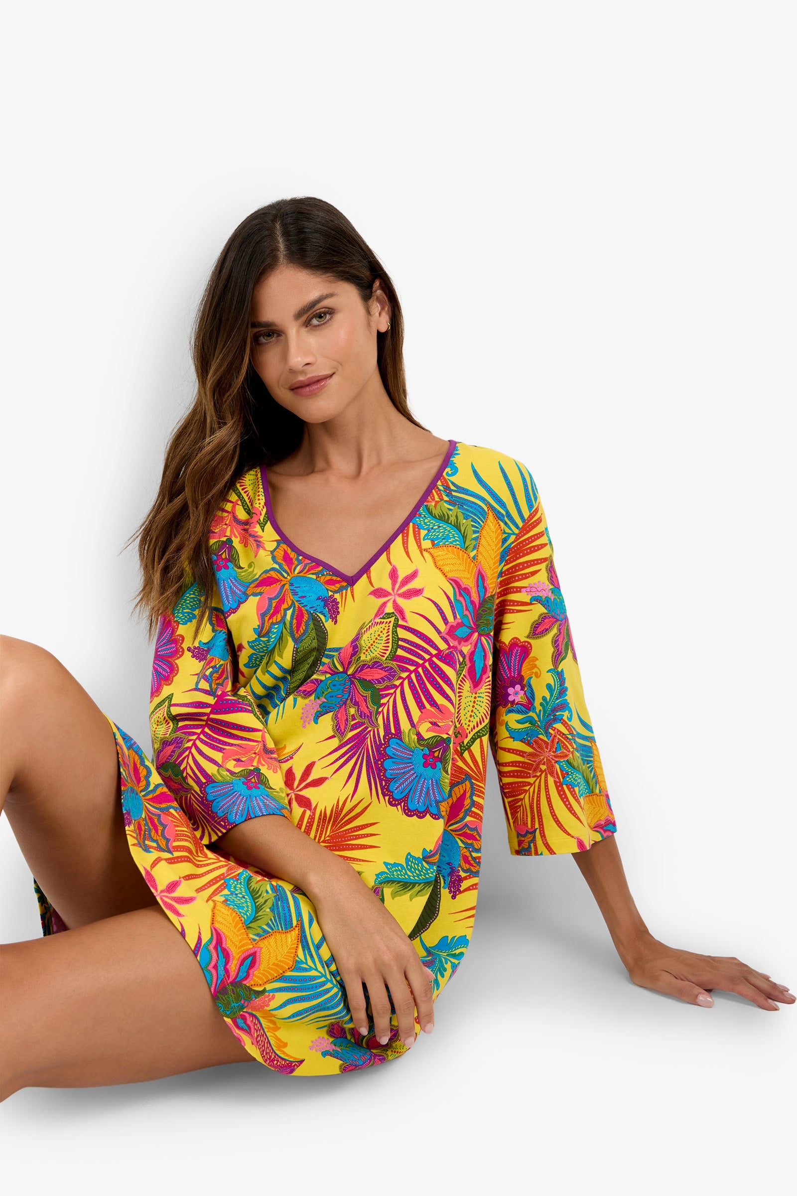 A woman wearing a yellow tropical-print Sleepshirt adorned with colorful flowers and leaves sits on the floor against a white background, looking at the camera with a relaxed expression.