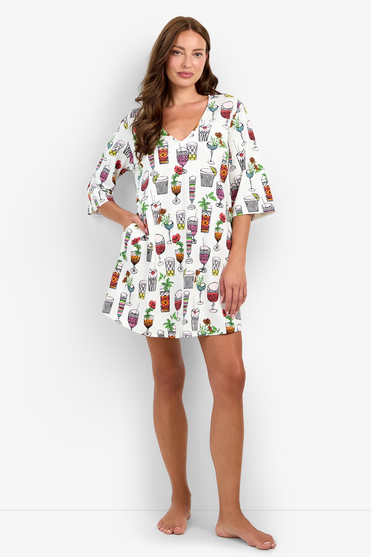 A woman stands barefoot against a plain white background, wearing the Sleepshirt—a white dress featuring colorful cocktail glass prints—with one hand in her pocket.
