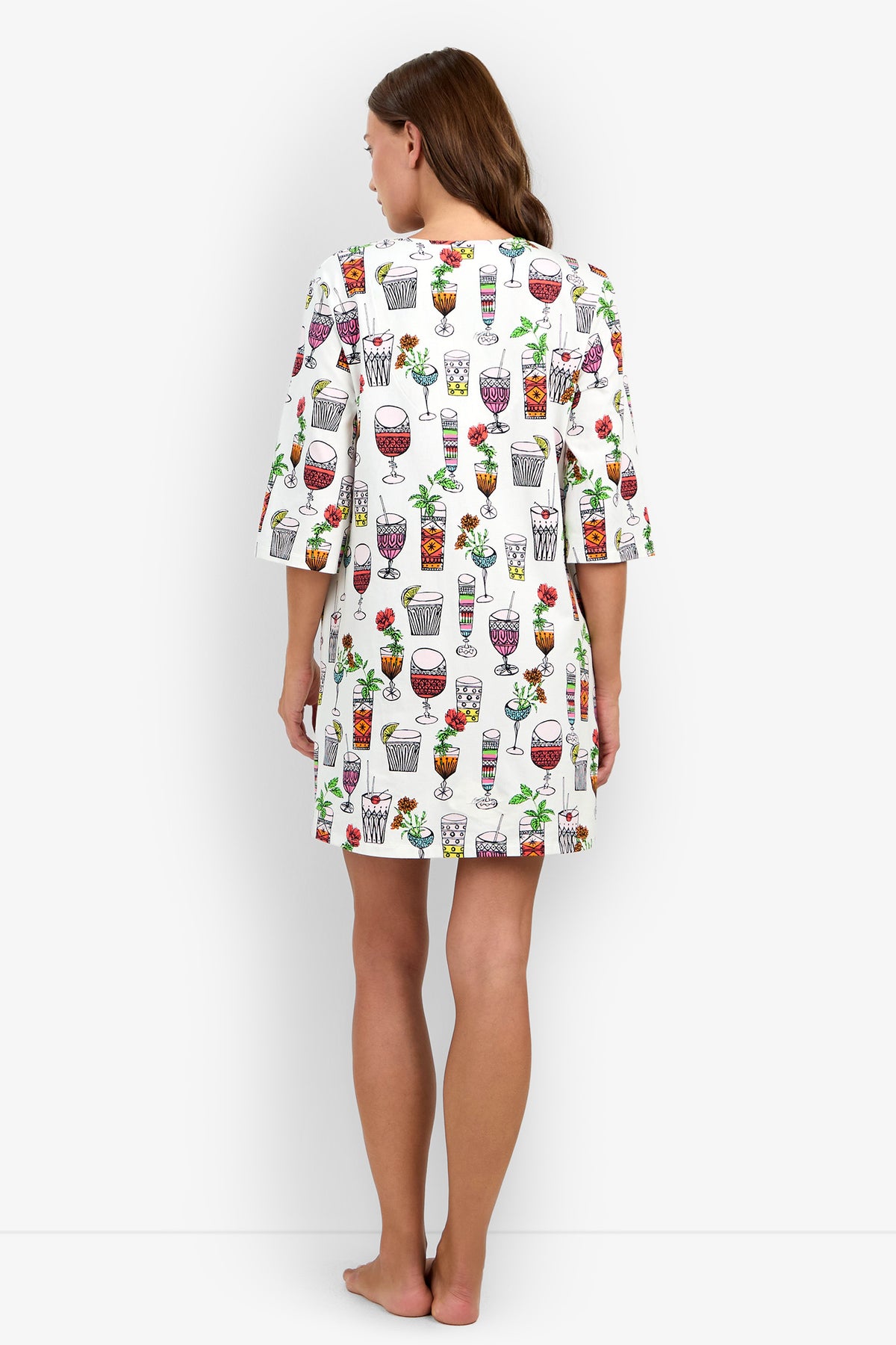 A woman stands barefoot facing away, wearing a Sleepshirt with colorful drink and fruit prints. The white Sleepshirt features elbow-length sleeves and falls above the knees. The background is plain white.