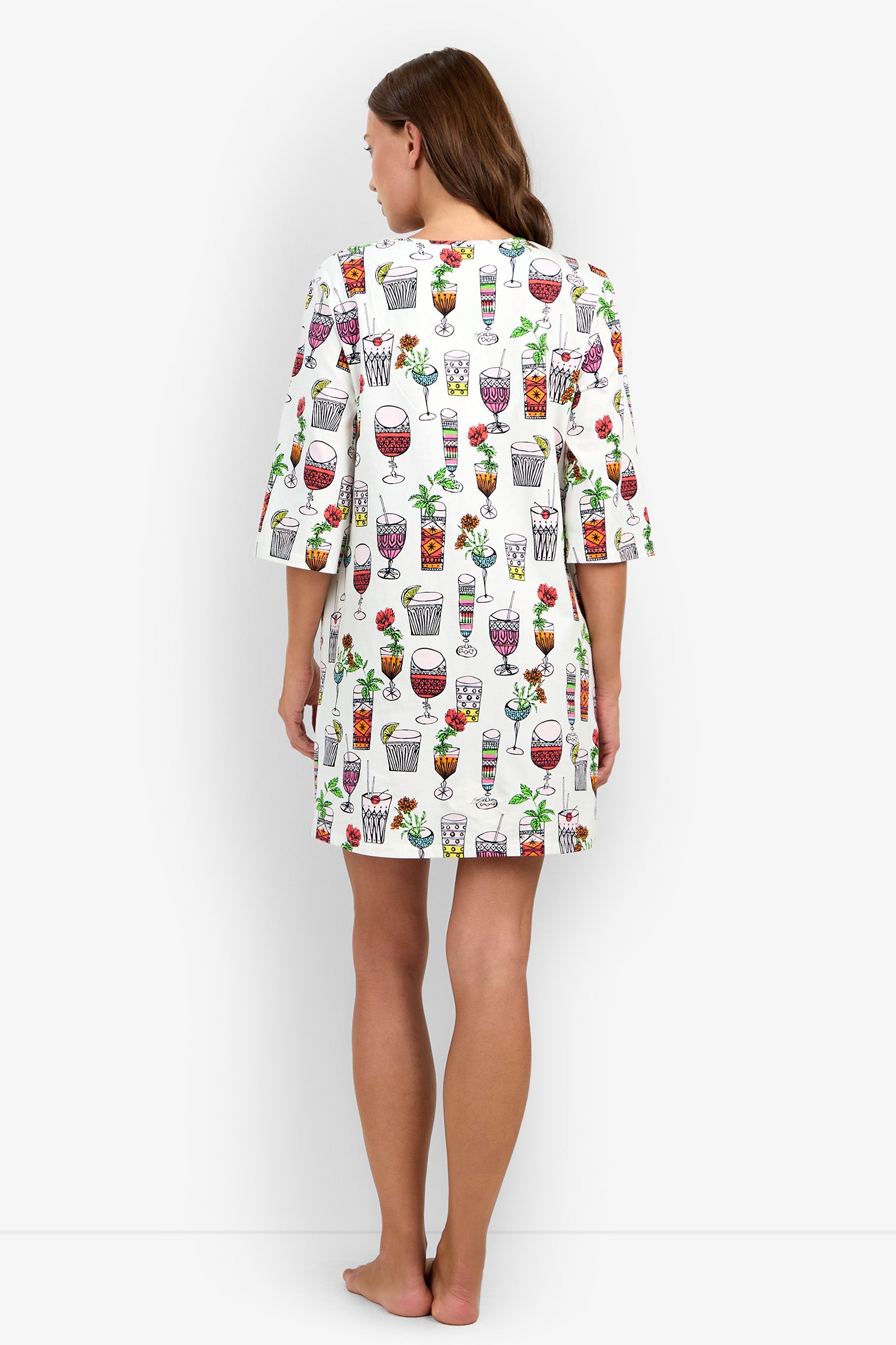 A woman stands barefoot facing away, wearing a Sleepshirt with colorful drink and fruit prints. The white Sleepshirt features elbow-length sleeves and falls above the knees. The background is plain white.