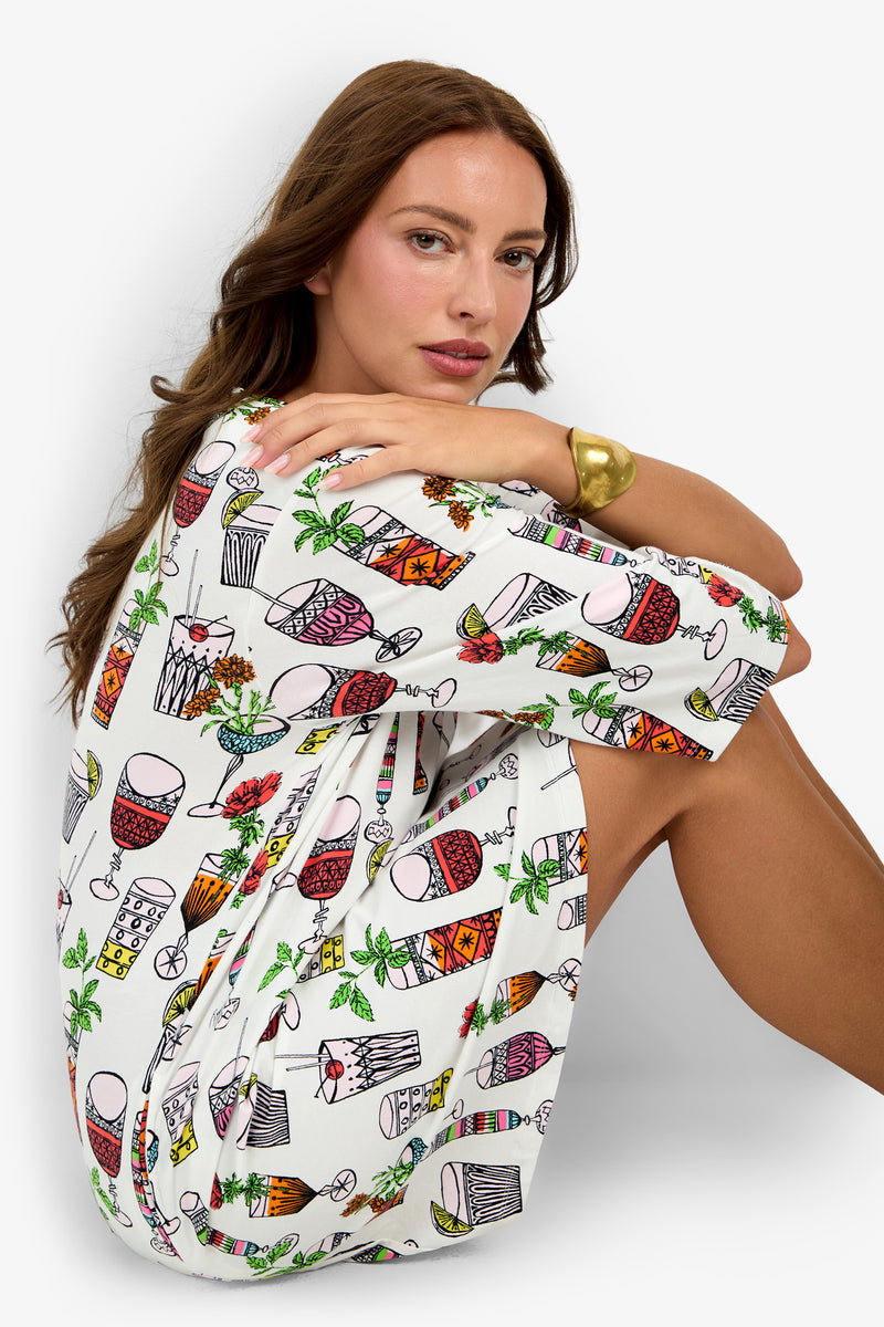 A woman with long brown hair sits with her knees up, wearing a white Sleepshirt adorned with colorful cocktail and drink illustrations. She wears a gold bangle on her wrist and looks toward the camera against a plain light background.