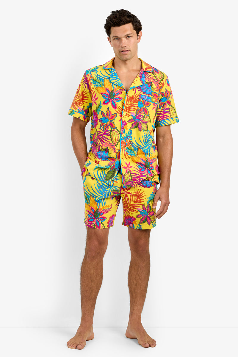 A man stands barefoot against a plain white background, wearing the Short Sleeve Boxer Pajamas—a bright yellow button-up shirt and matching shorts featuring a vibrant tropical floral pattern in red, blue, teal, and pink.
