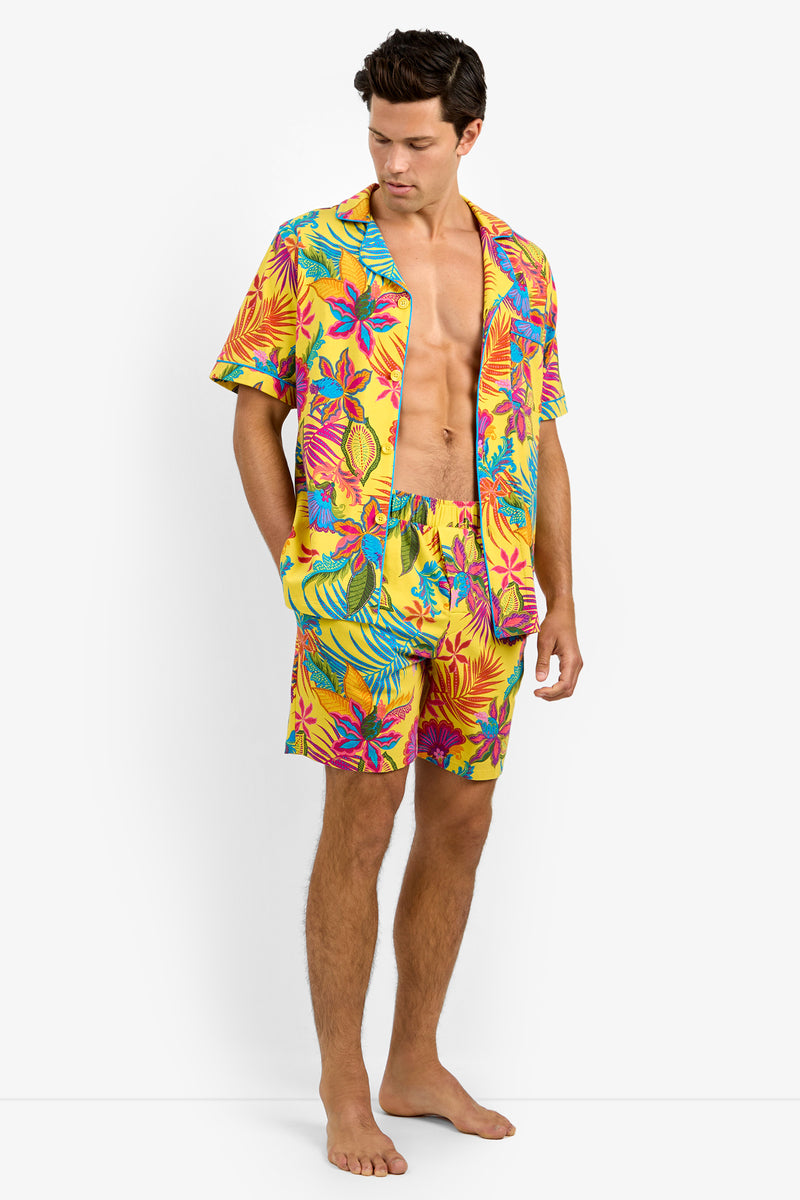 A man stands barefoot wearing Short Sleeve Boxer Pajamas in a bright yellow, pink, and blue tropical floral pattern. He looks down with his hands in his pockets.
