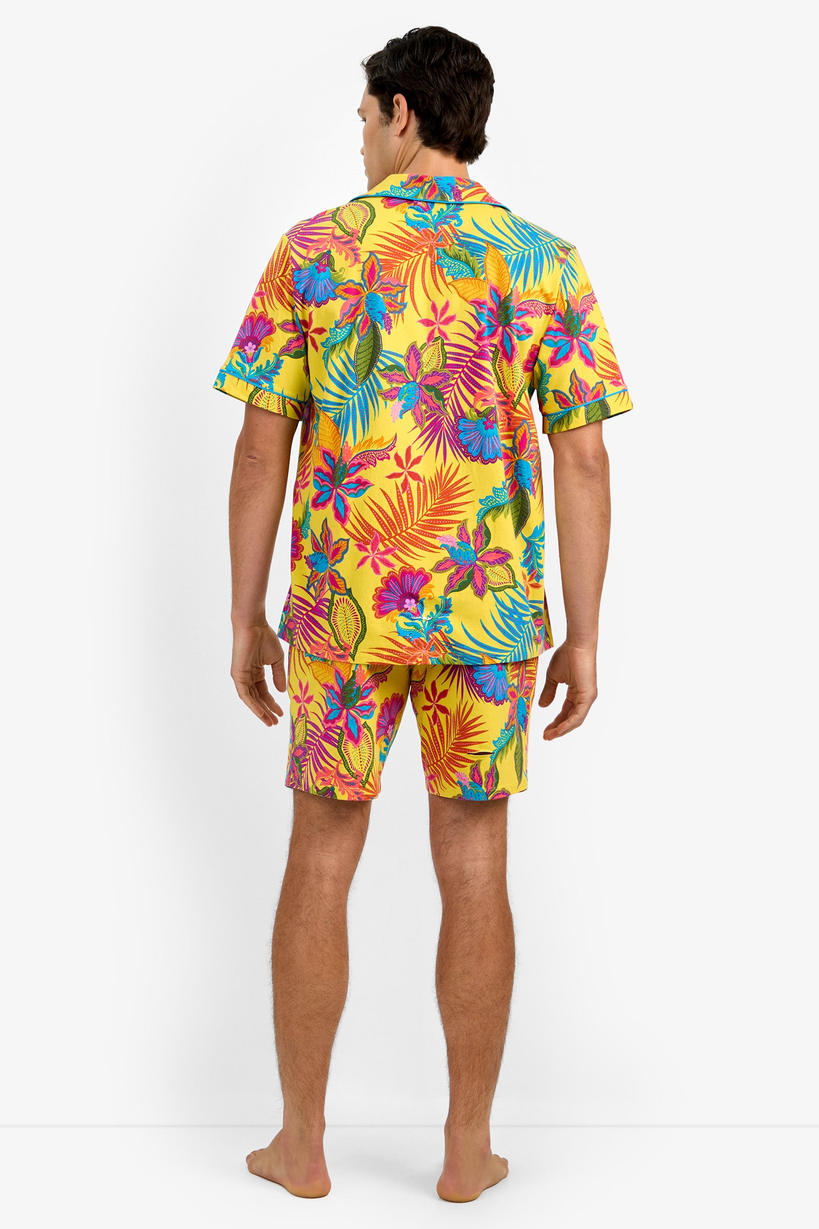 A man stands barefoot facing away, wearing Short Sleeve Boxer Pajamas featuring a matching yellow shirt and shorts with a vibrant tropical floral pattern of pink, blue, and orange leaves and flowers.