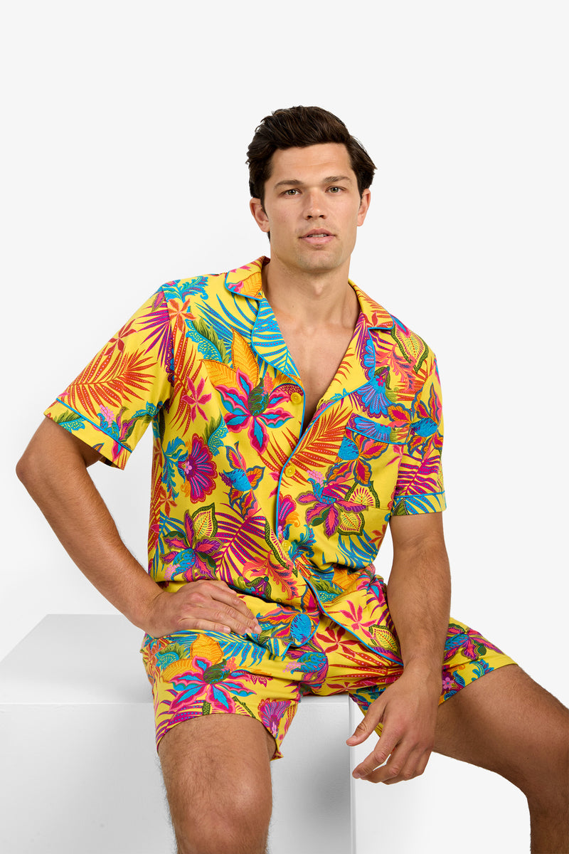 A man sits on a white block wearing Short Sleeve Boxer Pajamas with a bold tropical floral pattern in yellow, blue, pink, orange, and green.