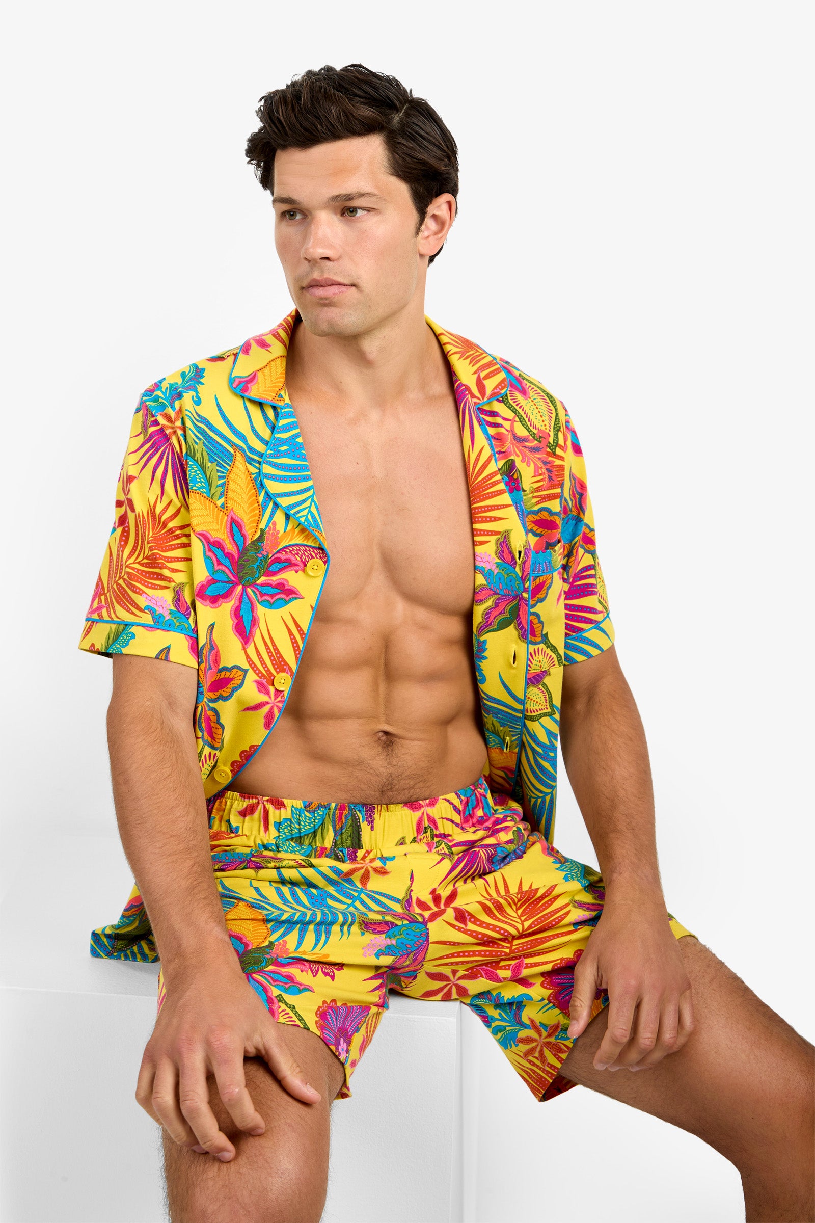 A man sits on a white block wearing Short Sleeve Boxer Pajamas, featuring a matching bright yellow set with a colorful tropical floral pattern. He looks slightly to the side with a neutral expression, revealing his chest and toned abs.
