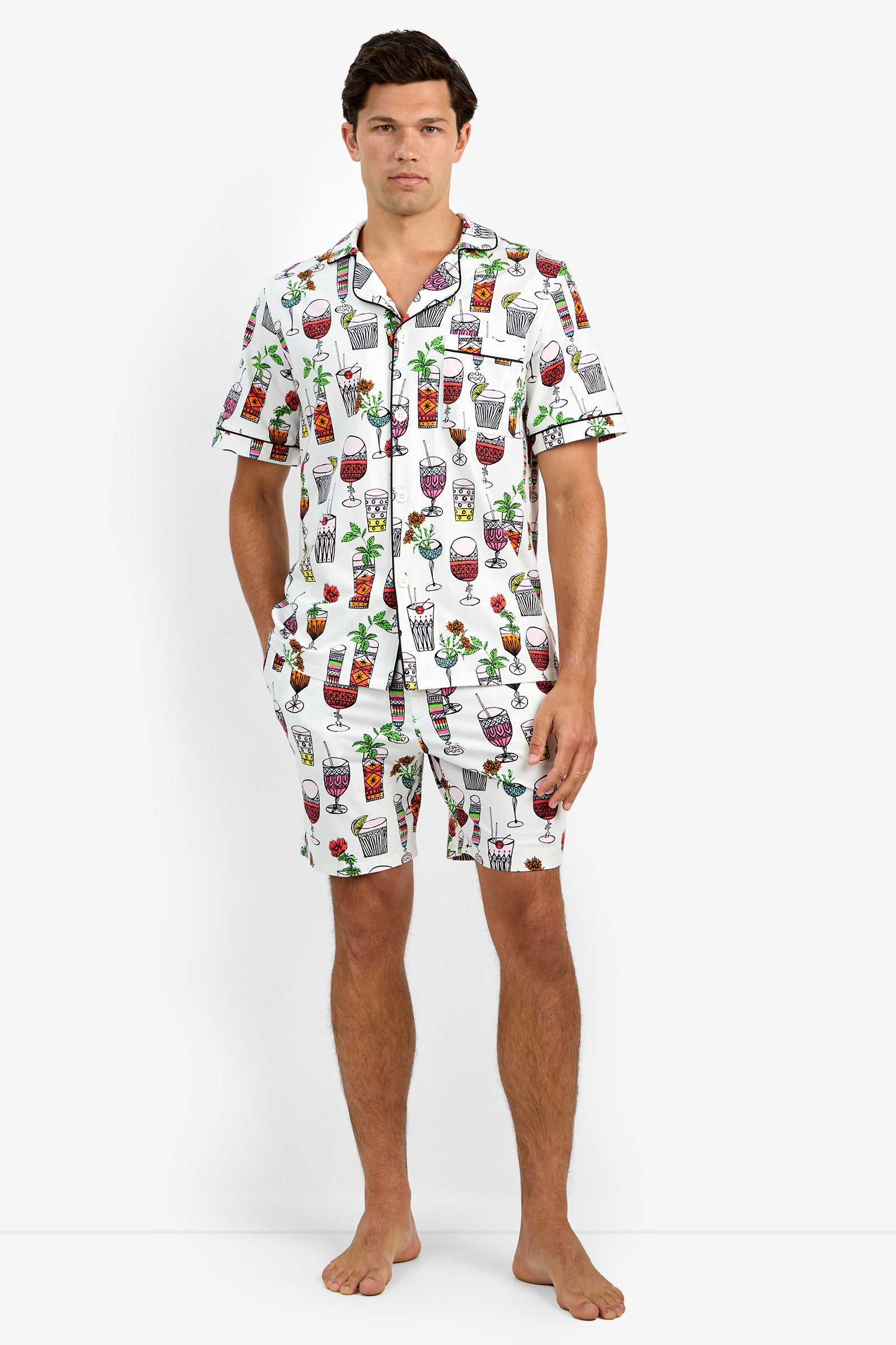 A man stands barefoot wearing Short Sleeve Boxer Pajamas in Happy Hour, featuring a colorful cocktail print. He poses with one hand in his pocket against a plain white background.