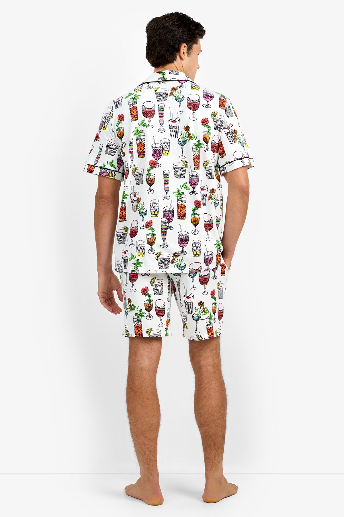 A man stands barefoot facing away, wearing Short Sleeve Boxer Pajamas featuring a matching button-up shirt and shorts with a colorful cocktail and drink glass pattern on a white background.