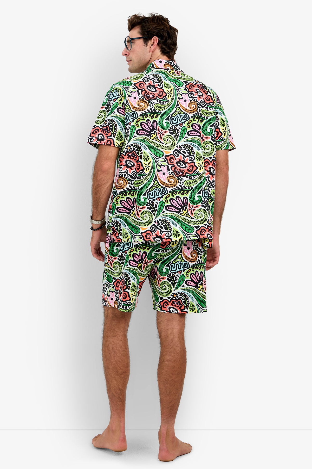 A barefoot man stands facing away, wearing the Short Sleeve Boxer Pajamas Set featuring a matching green, pink, and orange paisley pattern. He has short brown hair and glasses, with his hands relaxed by his sides.