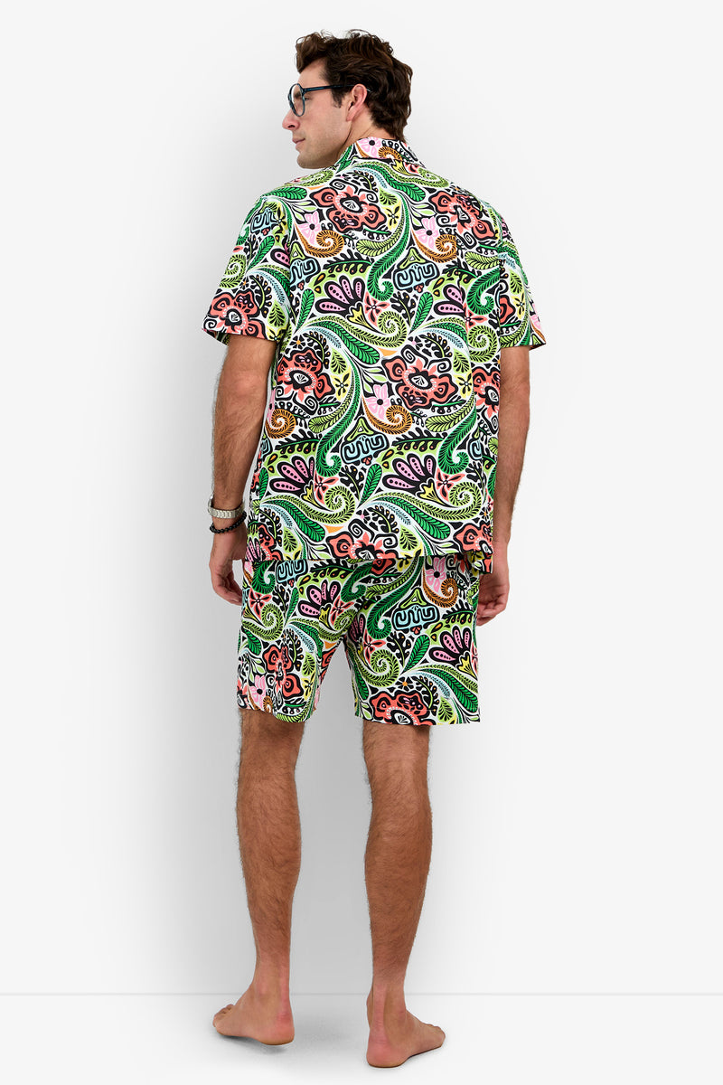 A barefoot man stands facing away, wearing the Short Sleeve Boxer Pajamas Set featuring a matching green, pink, and orange paisley pattern. He has short brown hair and glasses, with his hands relaxed by his sides.