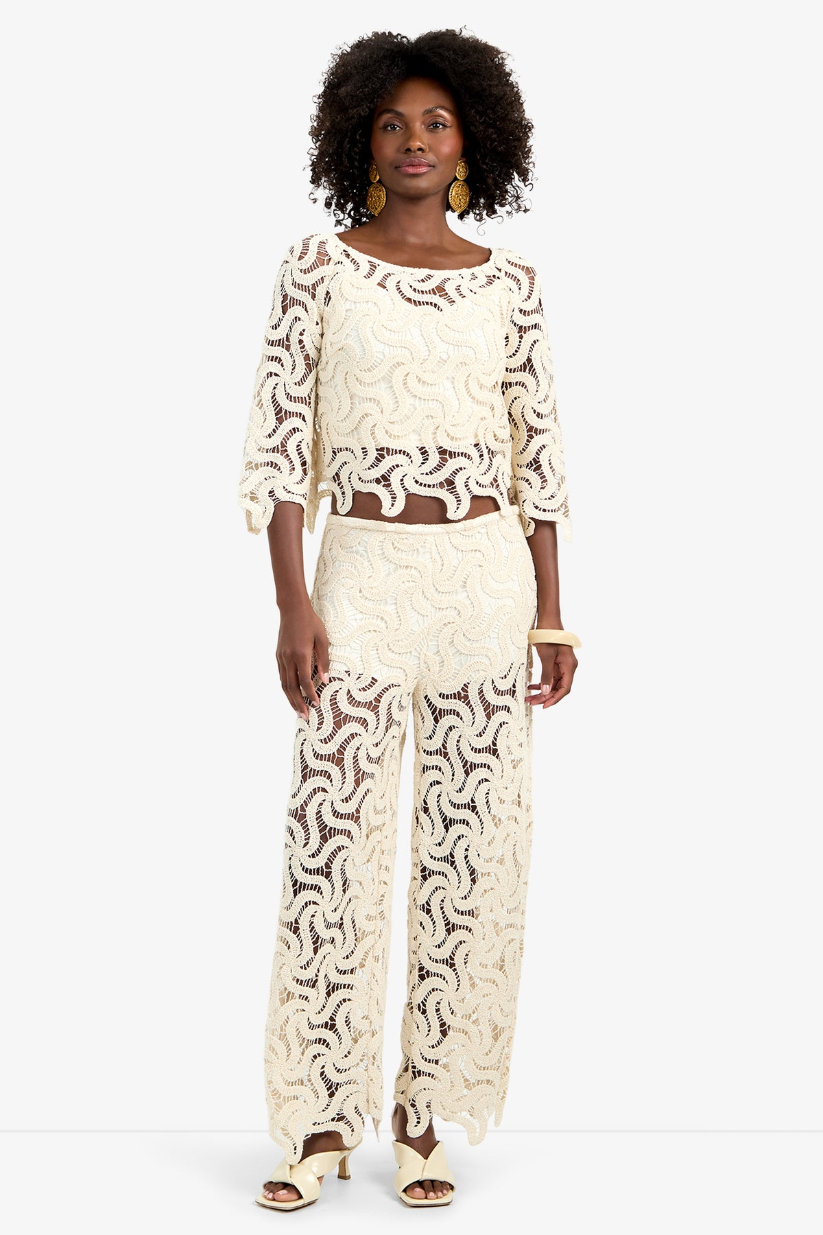 A woman with short curly hair, gold earrings, and heeled sandals stands facing forward in the Helen Top—a cream-colored, see-through lace top with a wavy pattern—paired with matching wide-leg pants.