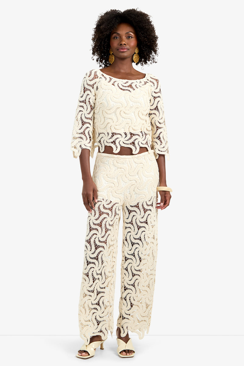 A woman with short curly hair, gold earrings, and heeled sandals stands facing forward in the Helen Top—a cream-colored, see-through lace top with a wavy pattern—paired with matching wide-leg pants.