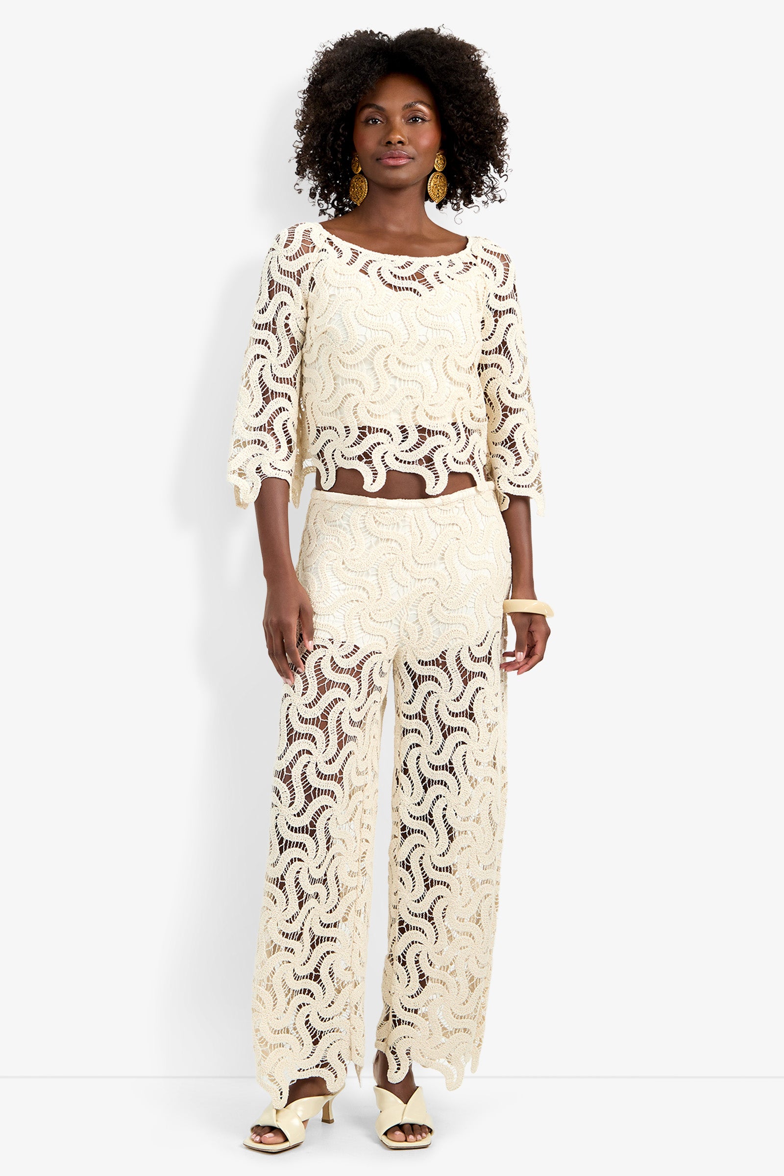 A Black woman models the Helen Top, a cream crochet crop top, paired with matching wide-leg pants, large gold earrings, and cream heeled sandals.