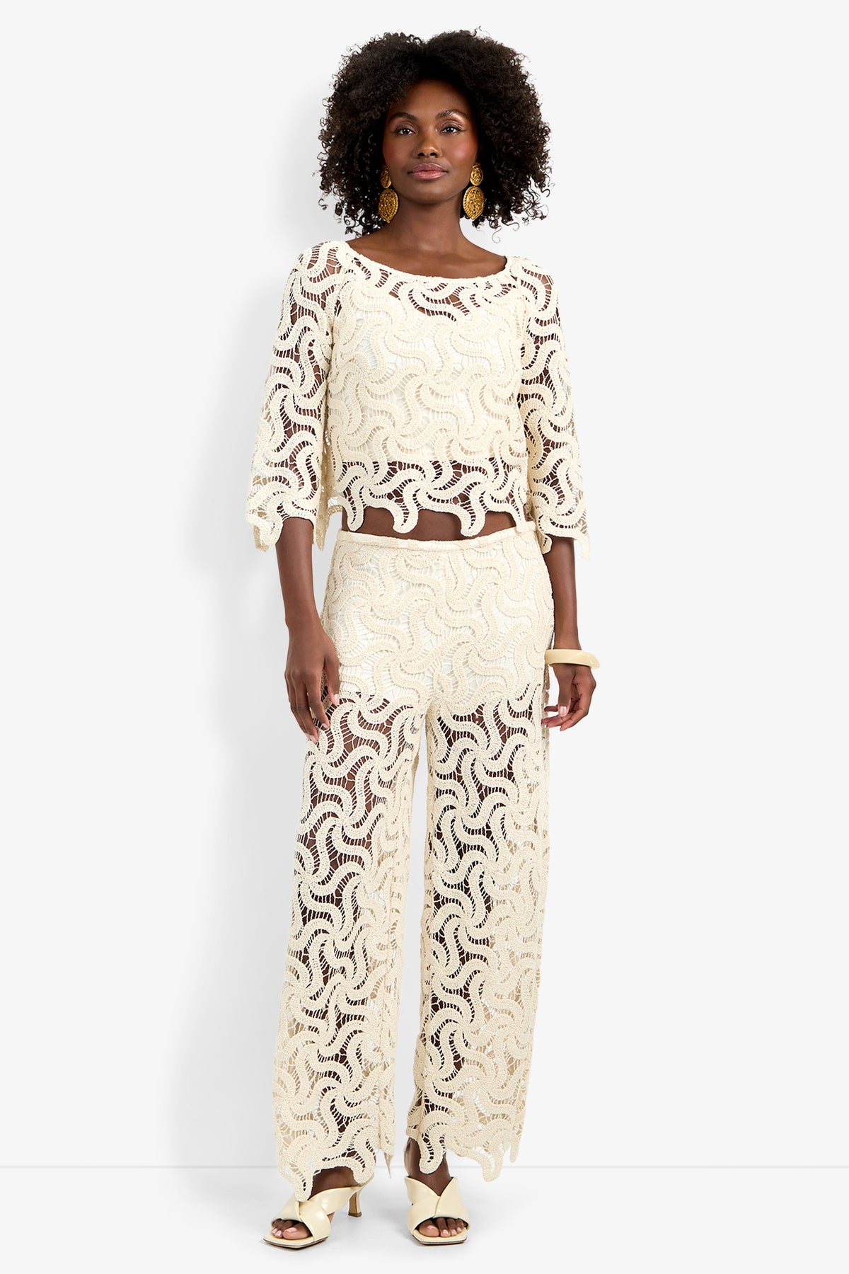 A woman stands against a white background wearing the Helen Top, a cream-colored lace two-piece set with loose three-quarter sleeves and wide-leg pants. She has curly hair, gold earrings, and cream heeled sandals.