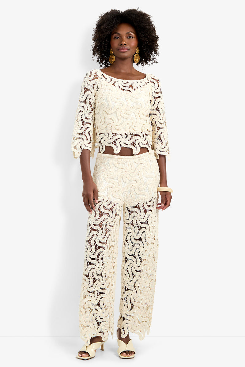 A woman stands against a white background wearing the Helen Top, a cream-colored lace two-piece set with loose three-quarter sleeves and wide-leg pants. She has curly hair, gold earrings, and cream heeled sandals.