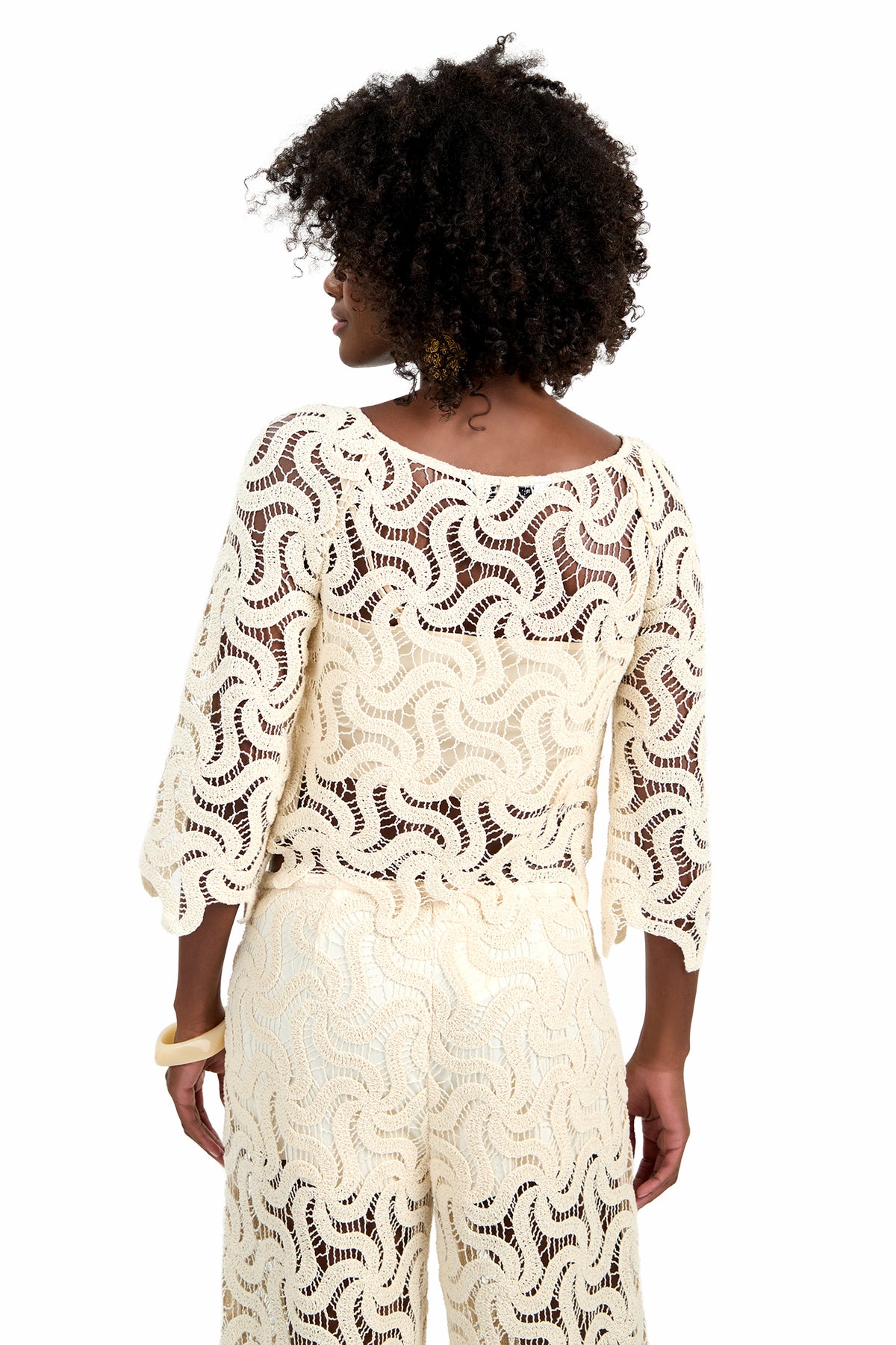 Woman with curly hair faces away, wearing the Helen Top in cream—an open-knit crochet design paired with matching pants. The white background highlights the texture and style of the outfit.