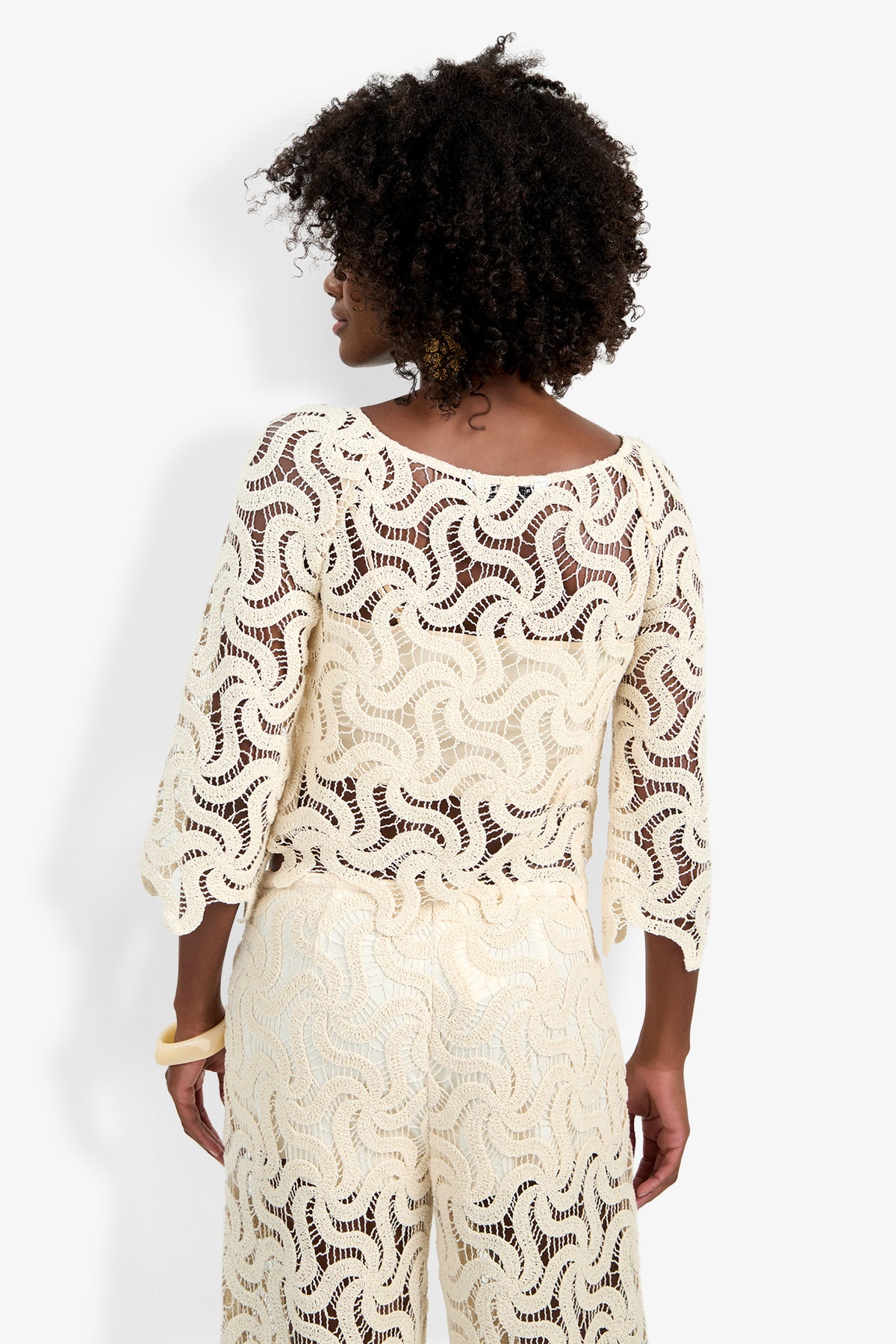 A woman with curly hair, facing away, wears the cream-colored Helen Top—a long-sleeve crocheted piece with a wavy, open-knit pattern—paired with matching pants and a light bangle on her left wrist, set against a plain white background.