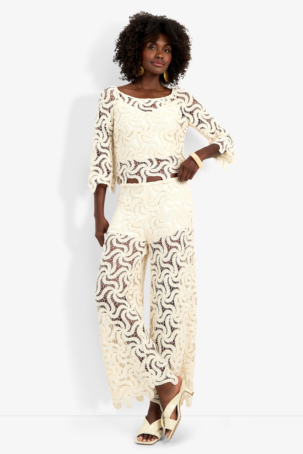 A woman wearing the Helen Top—a cream crochet long-sleeve piece—with matching wide-leg pants, gold hoop earrings, a bangle, and gold sandals stands against a plain white background.