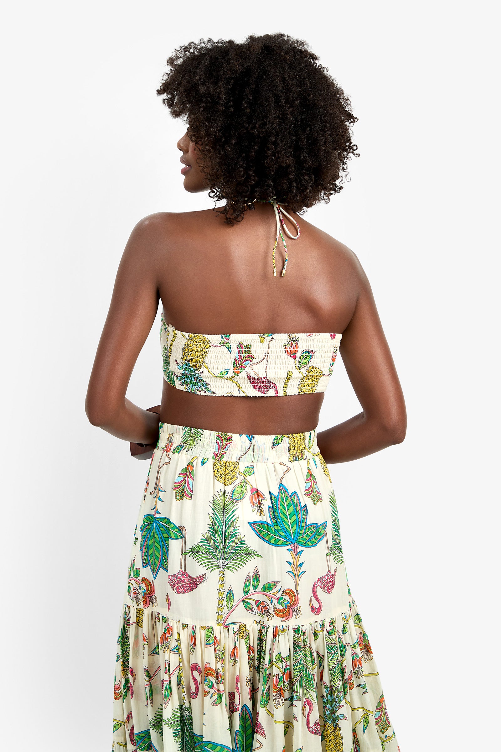 A woman with curly hair faces away, showing the back details of the Marisalle Top—a strapless, tropical-print piece with leaves, flamingos, and fruit patterns—against a white background.
