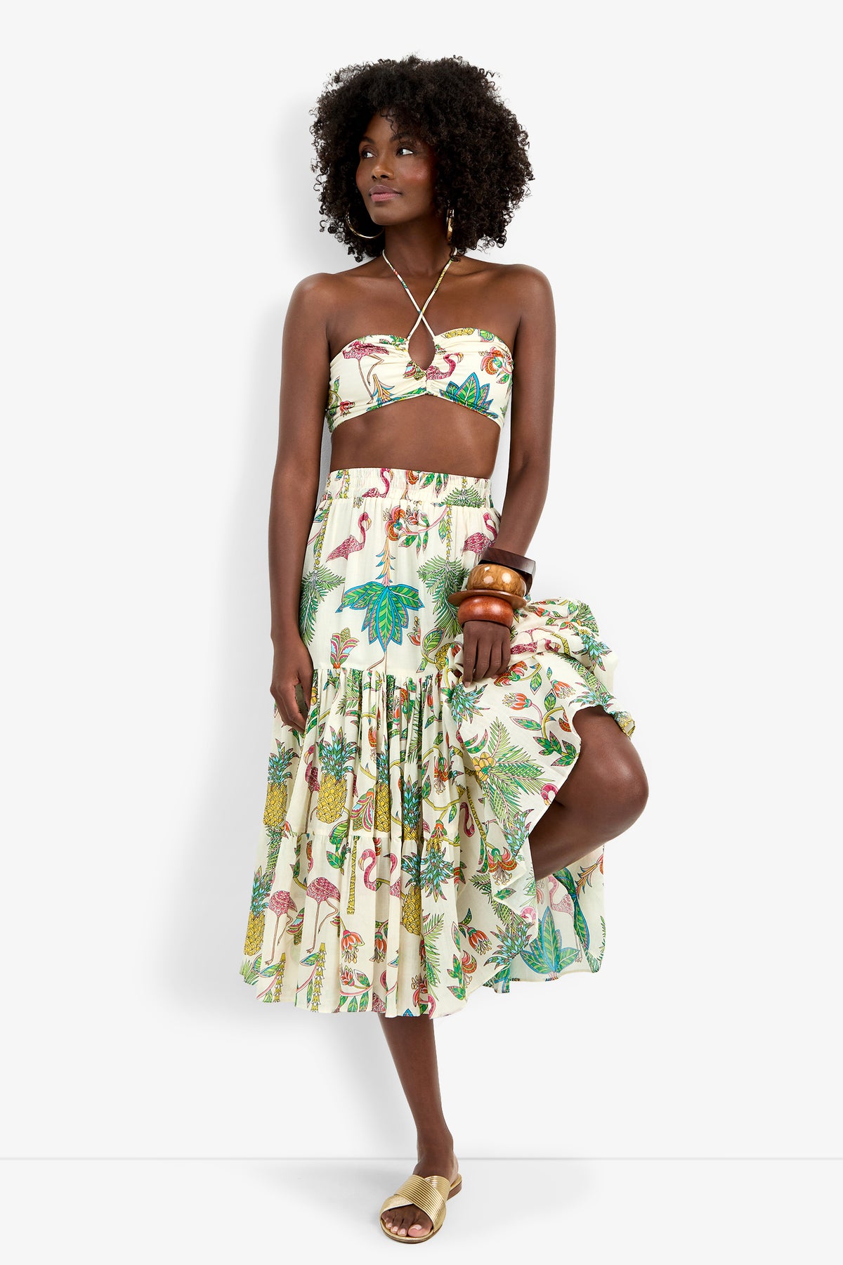 A woman in the Marisalle Top and matching tropical print midi skirt poses with one knee raised, accessorizing with gold sandals, wooden bangles, and a necklace against a white background.