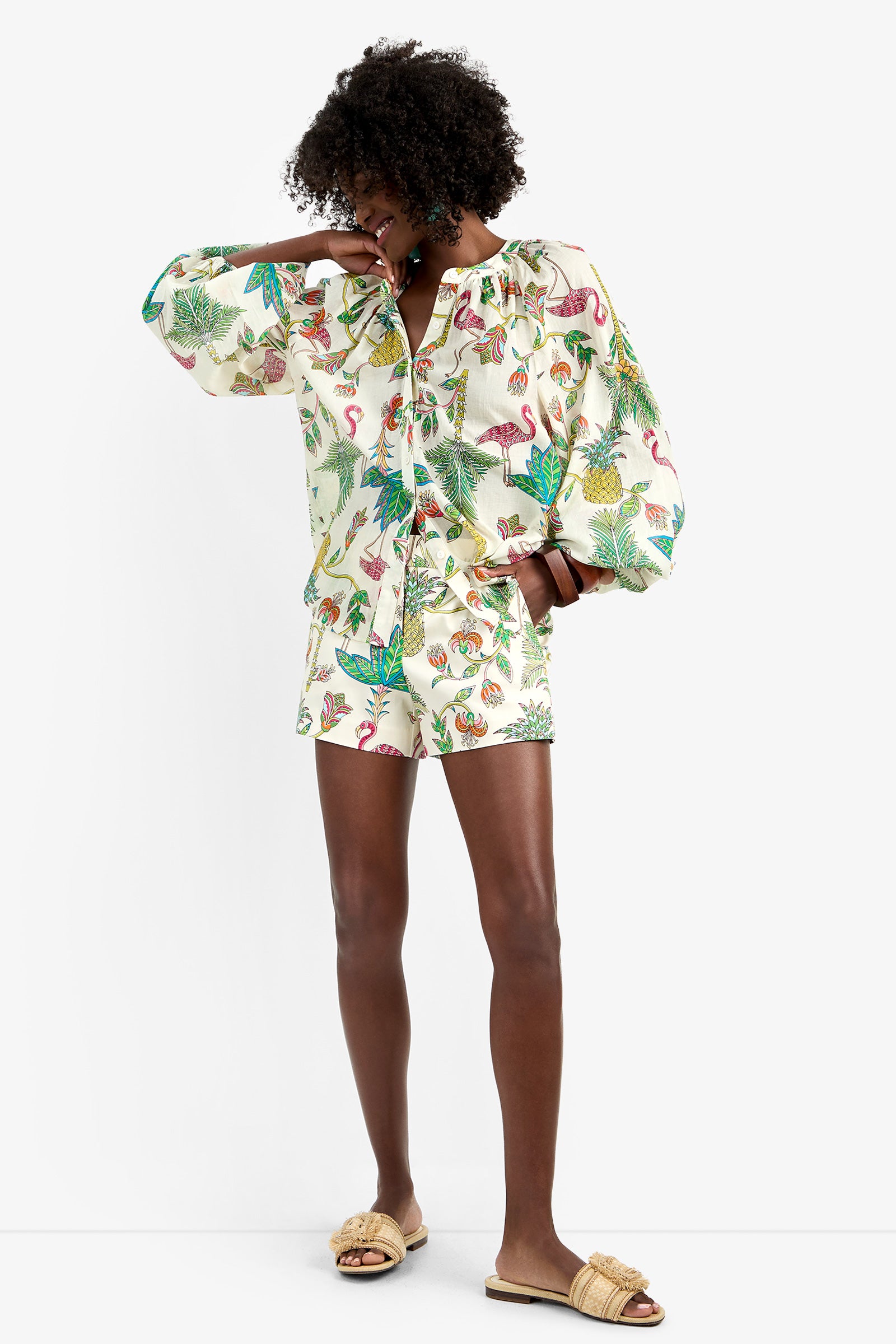 A person smiles in the NORRY TOP, a matching set with tropical flamingo and leaf prints made from 100% cotton. They pair it with tan slide sandals, raising one arm to partially cover their face against a plain white background.