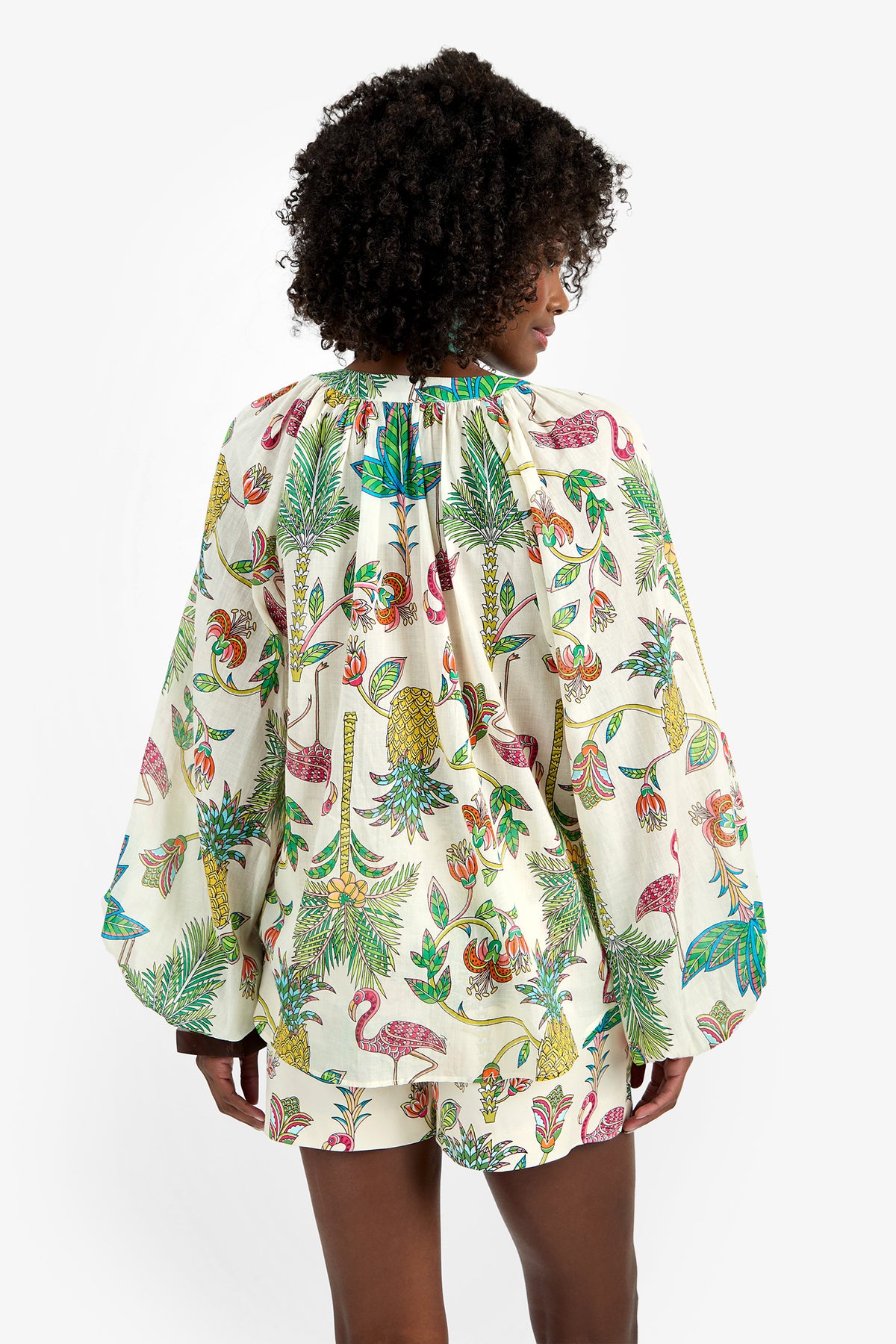 A person with curly hair, seen from behind, wears the NORRY TOP—100% cotton, long-sleeved and loose—with a colorful tropical print of plants, birds, and fruit on a white background.