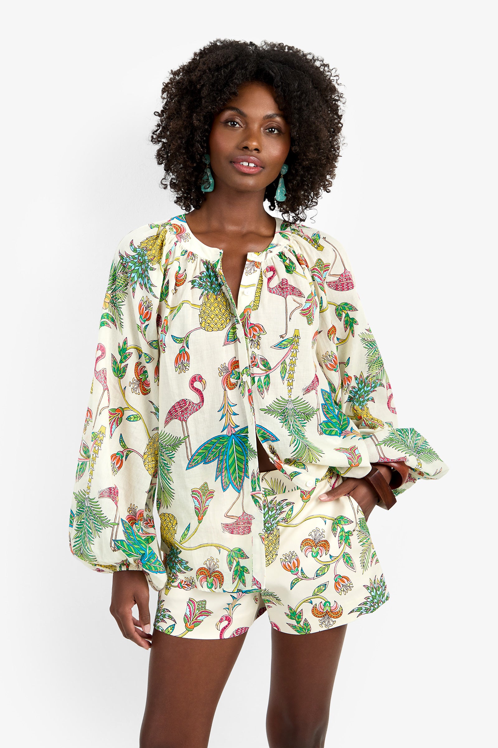 A woman with curly hair wears the NORRY TOP, a loose, long-sleeved cotton blouse with a vibrant tropical print of birds, pineapples, and leaves, paired with matching shorts against a white background.