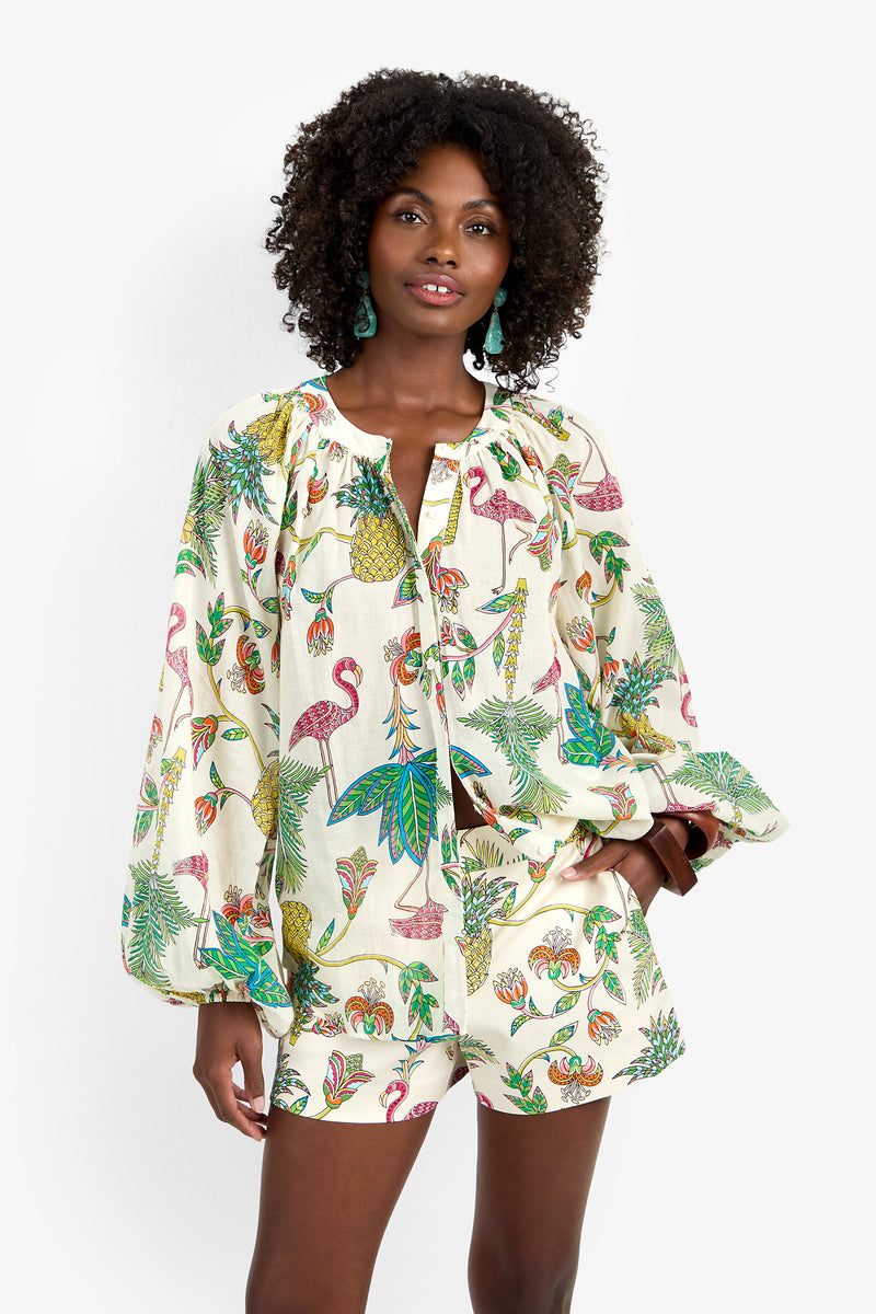 A woman with curly hair wears the NORRY TOP, a loose, long-sleeved cotton blouse with a vibrant tropical print of birds, pineapples, and leaves, paired with matching shorts against a white background.