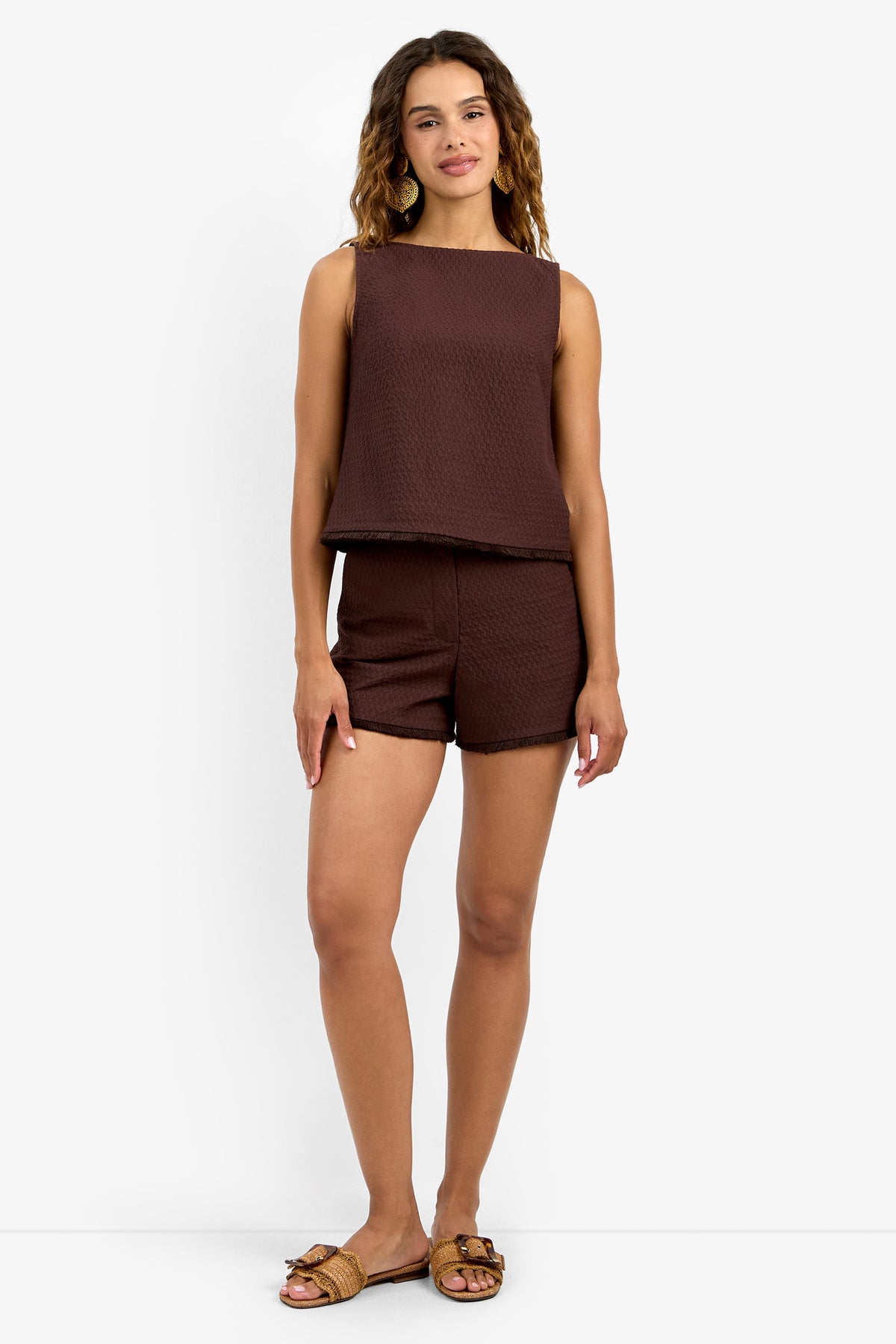 A woman with wavy hair and a relaxed smile stands against a white background, wearing the sleeveless brown textured Port Top with matching brown shorts and brown slide sandals.