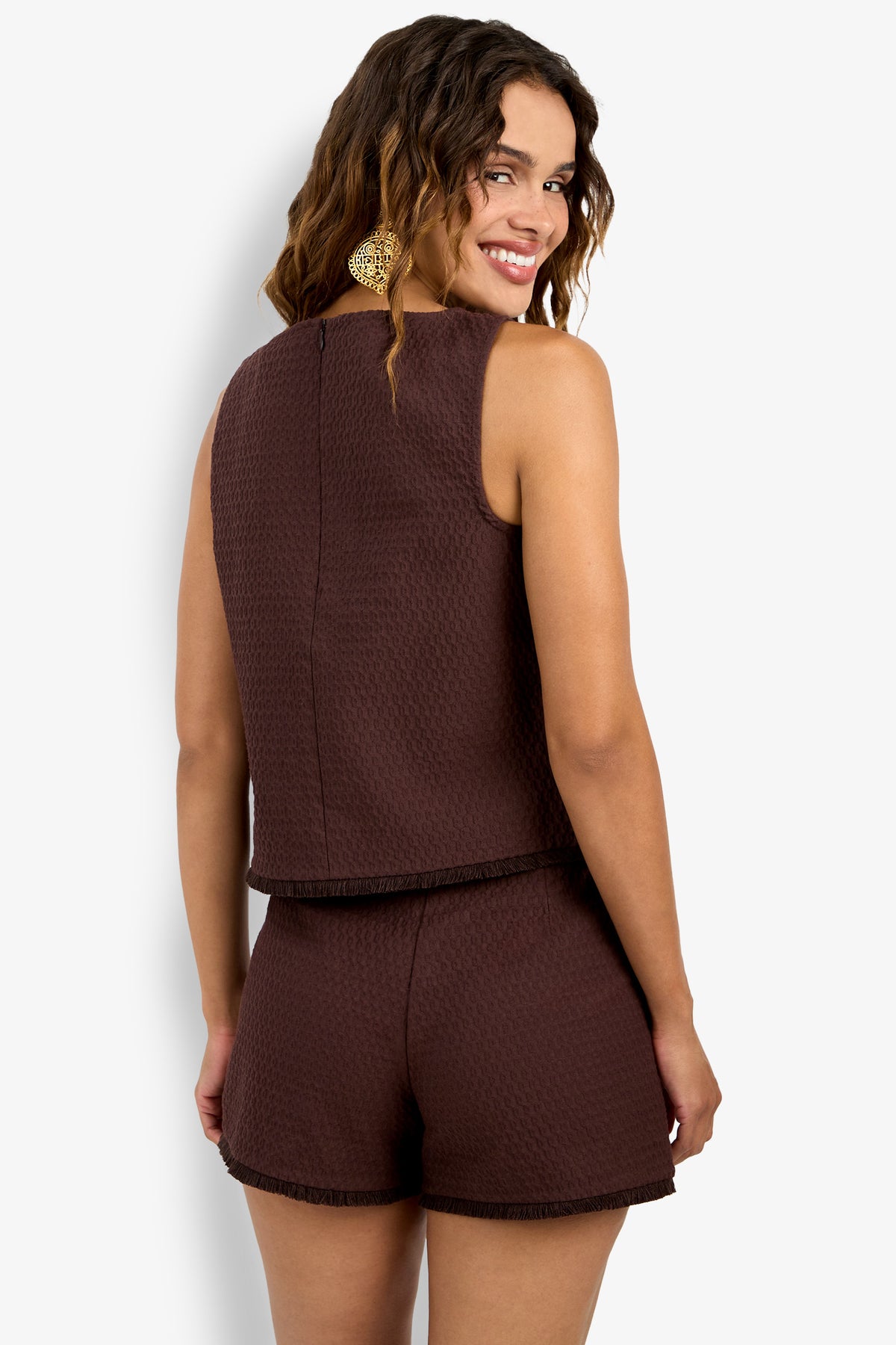 Woman with wavy brown hair smiles over her shoulder, wearing the Port Top— a sleeveless, textured dark brown top with fringe trim and matching shorts. She accessorizes with large earrings against a plain white background.