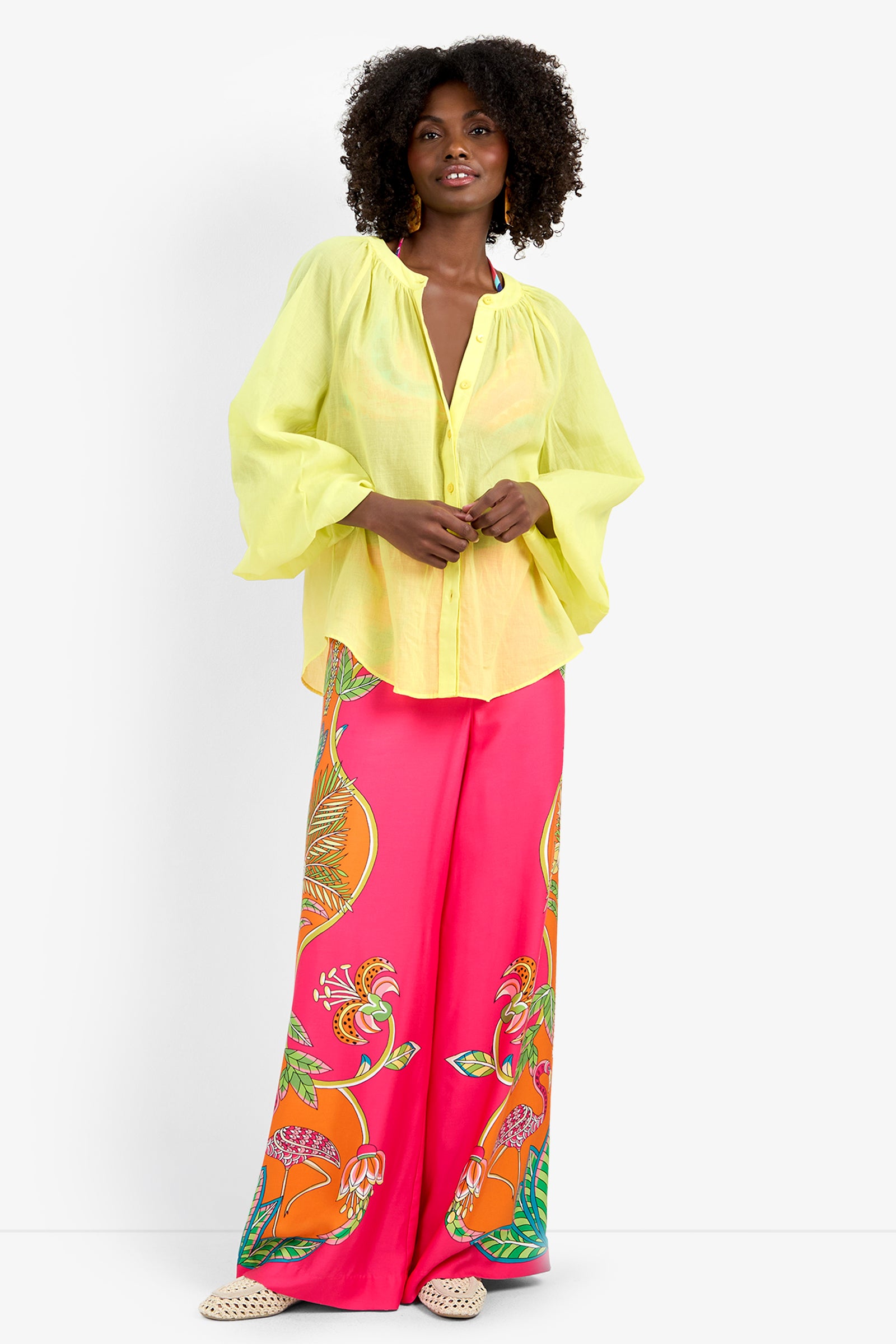 A woman with curly hair, gold earrings, and woven sandals stands against a white background wearing the NORRY TOP— a loose, sheer yellow blouse— paired with wide-leg pink pants featuring bold tropical prints in 100% cotton.