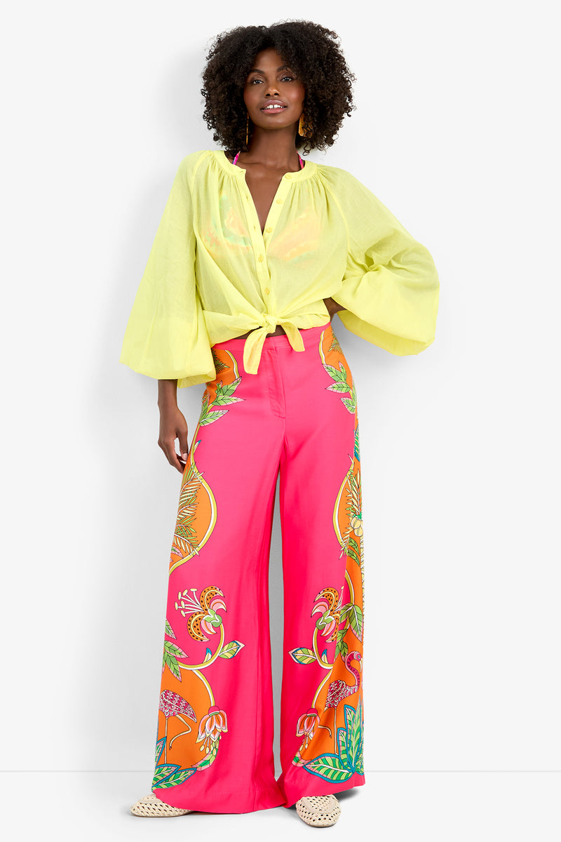 A woman stands smiling in the Norry Top, a yellow long-sleeved button-up blouse with a tie front, paired with wide-legged, high-waisted pink pants featuring bold floral patterns and light woven shoes.