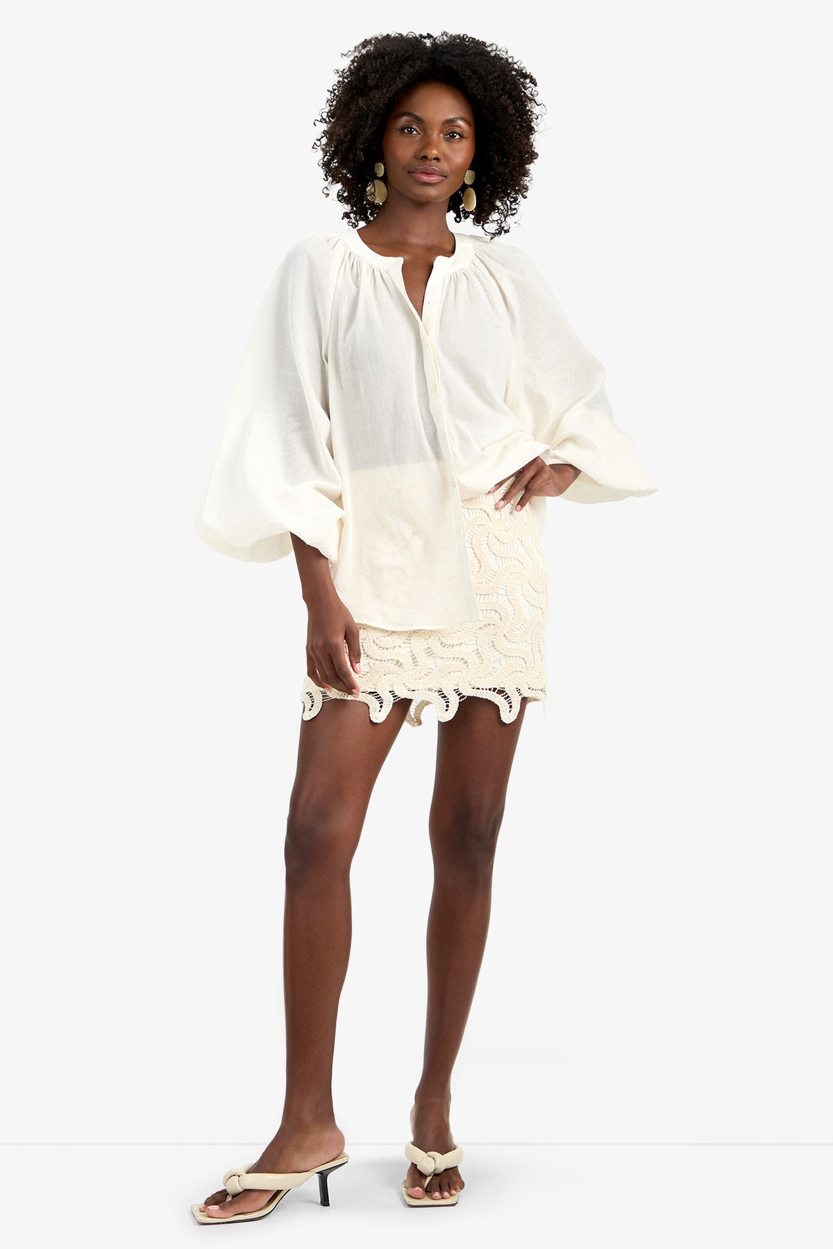 A woman poses against a plain background in the NORRY TOP—a loose, long-sleeved 100% cotton blouse in Bonita Beige with gathered cuffs—styled with a cream mini skirt, white heeled sandals, and statement earrings.