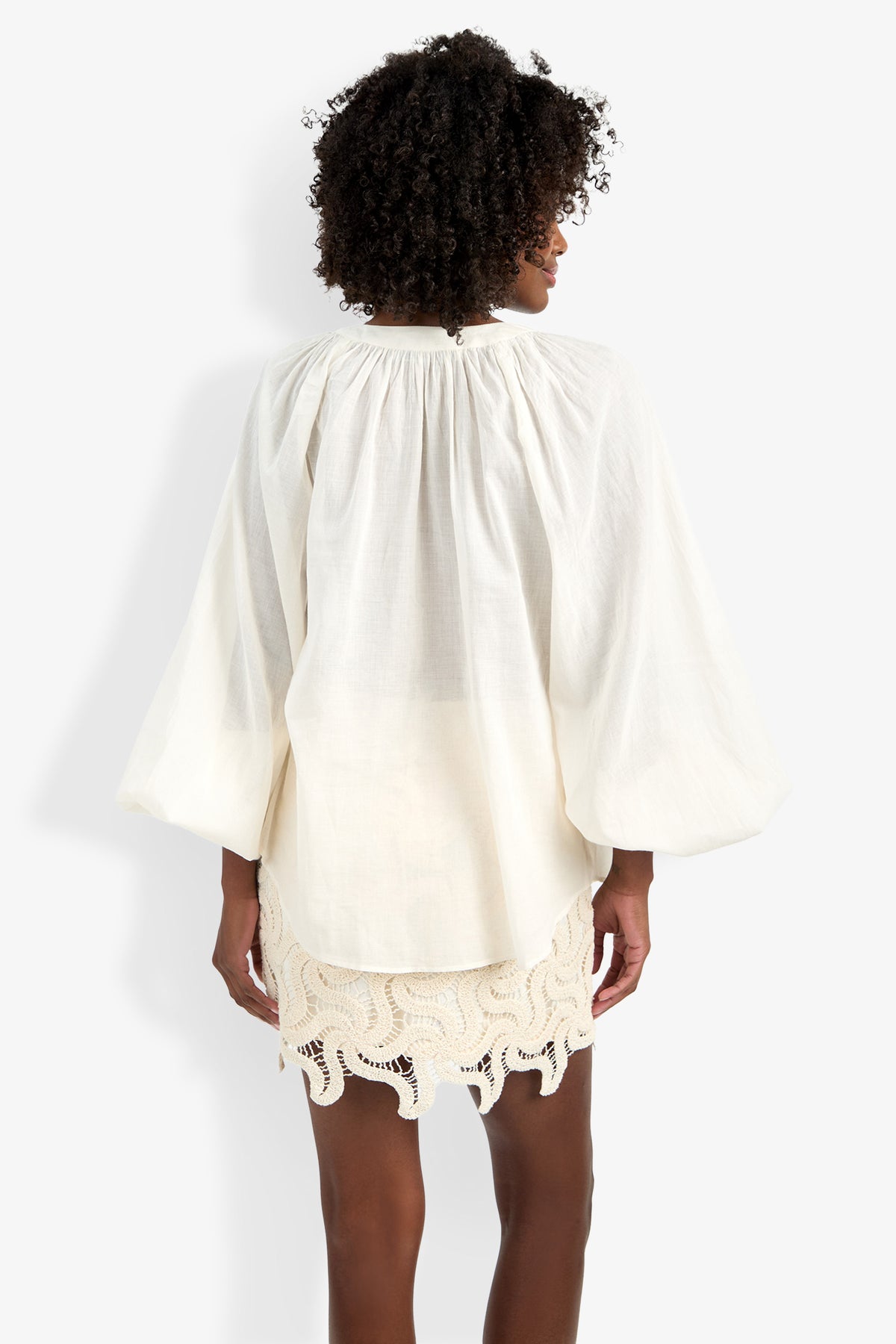 A woman with curly hair, seen from behind against a white background, wears the Norry Top—a loose, long-sleeved cream blouse—paired with a skirt featuring a lacy, scalloped hem.
