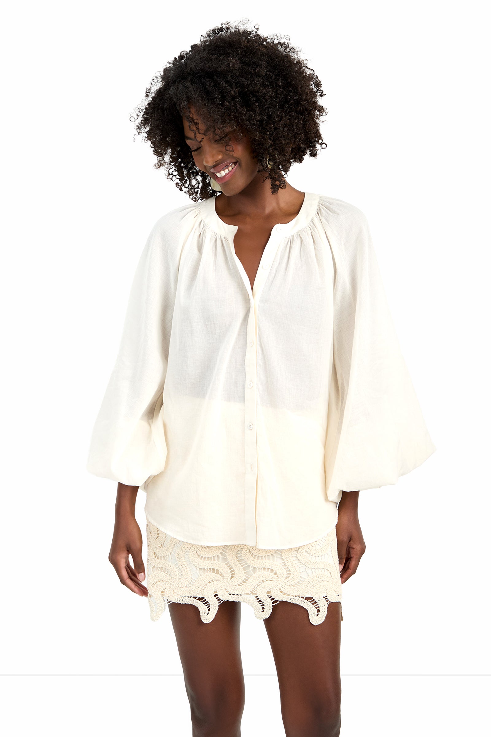 A smiling woman with curly hair wears the NORRY TOP, a loose, cream-colored cotton blouse with billowy sleeves, and a short cream lace skirt, standing against a white background.