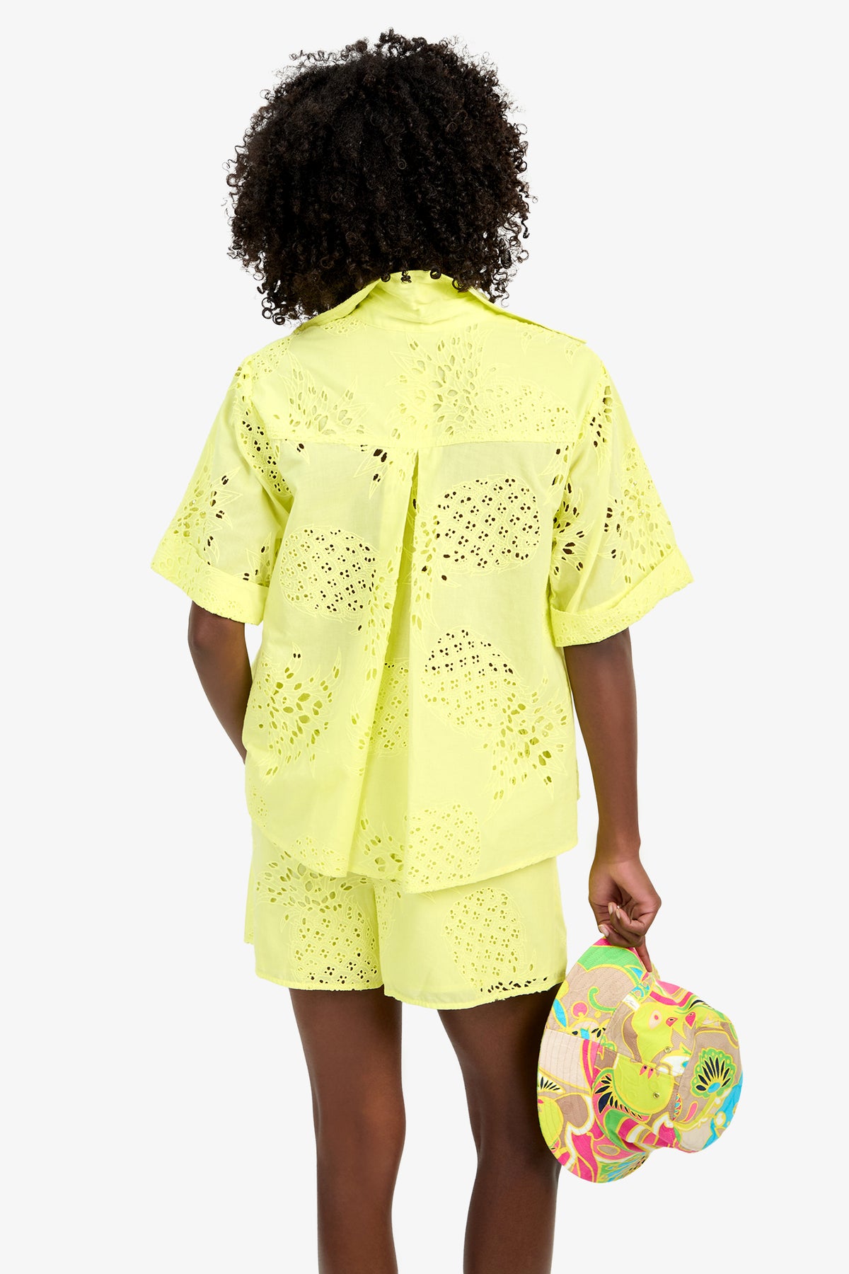 A person with curly hair, seen from behind, wears the TRIENNALE 2 TOP—a light yellow 100% cotton eyelet shirt with matching shorts—and holds a colorful tropical-patterned hat in their right hand against a plain white background.