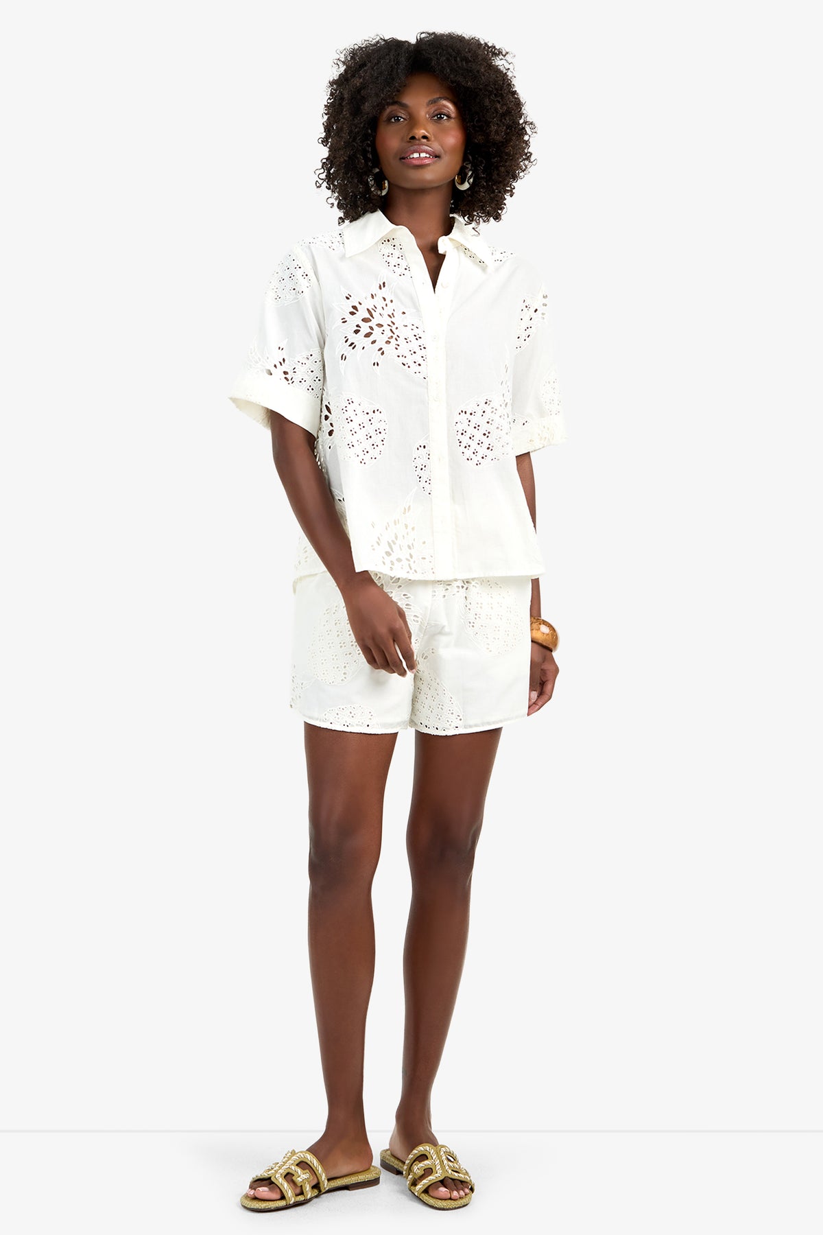 A woman with curly hair smiles, wearing the TRIENNALE 2 TOP—a 100% white cotton eyelet short-sleeve button-up shirt—paired with matching shorts, gold hoop earrings, a bracelet, and beige sandals with gold accents.
