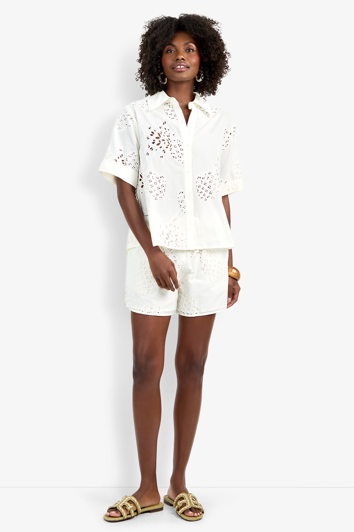 A woman with curly hair wears the white, short-sleeved Triennale Top with matching eyelet shorts, gold hoop earrings, a gold bracelet, and beige sandals with gold accents. The background is plain white.