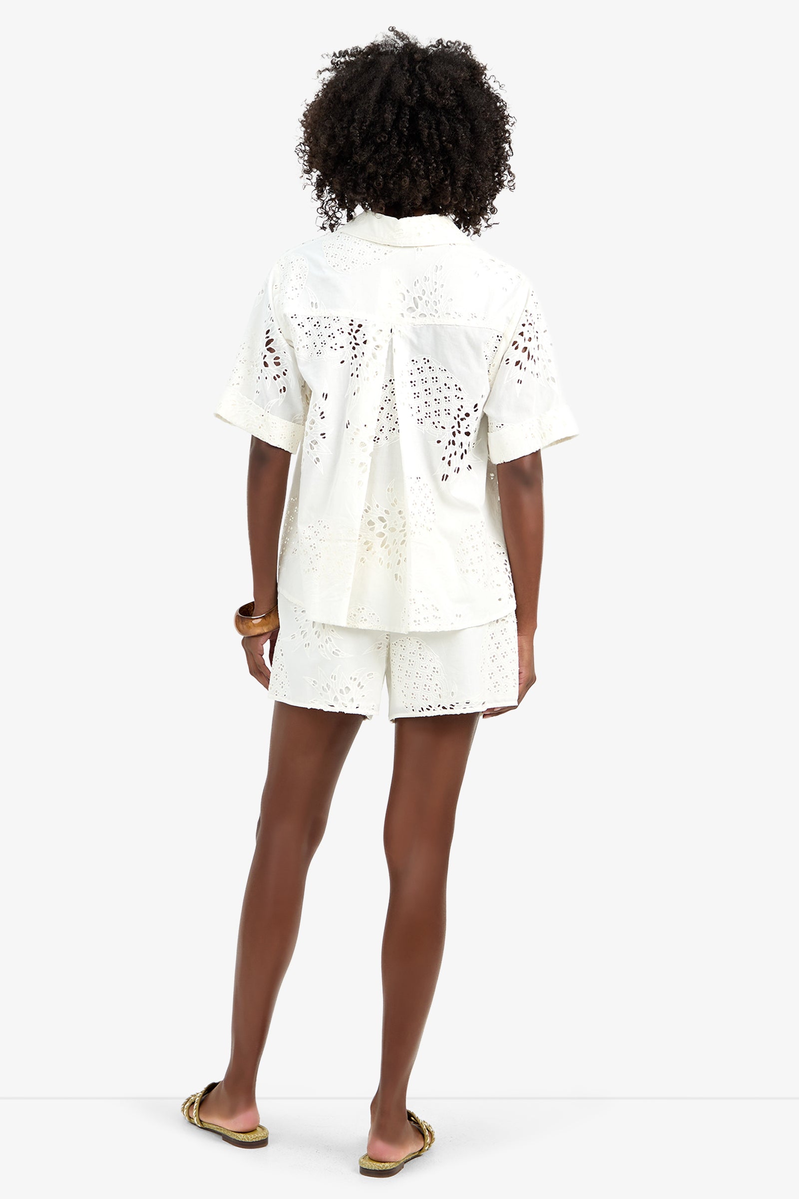 A person with curly hair, seen from the back, wears the TRIENNALE 2 TOP— a white short-sleeve 100% cotton eyelet shirt—paired with matching white eyelet shorts, a brown bracelet, and sandals, standing against a plain white background.