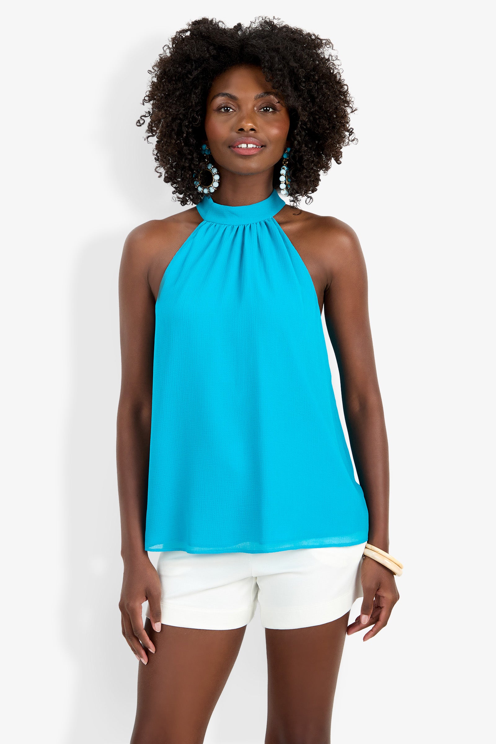 A woman models the Kissimmee Top, a bright blue halter-neck sleeveless style, paired with white shorts and accessorized with blue earrings and a bracelet against a plain white background.