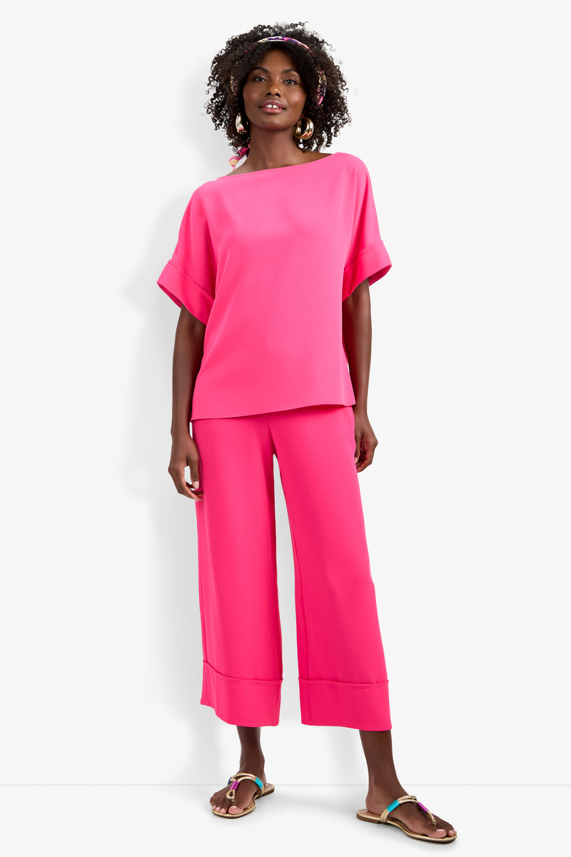 A woman with curly hair and hoop earrings wears the Sofie Top, a loose-fitting bright pink short-sleeve top, paired with matching wide-leg pants and colorful flat sandals against a white background.