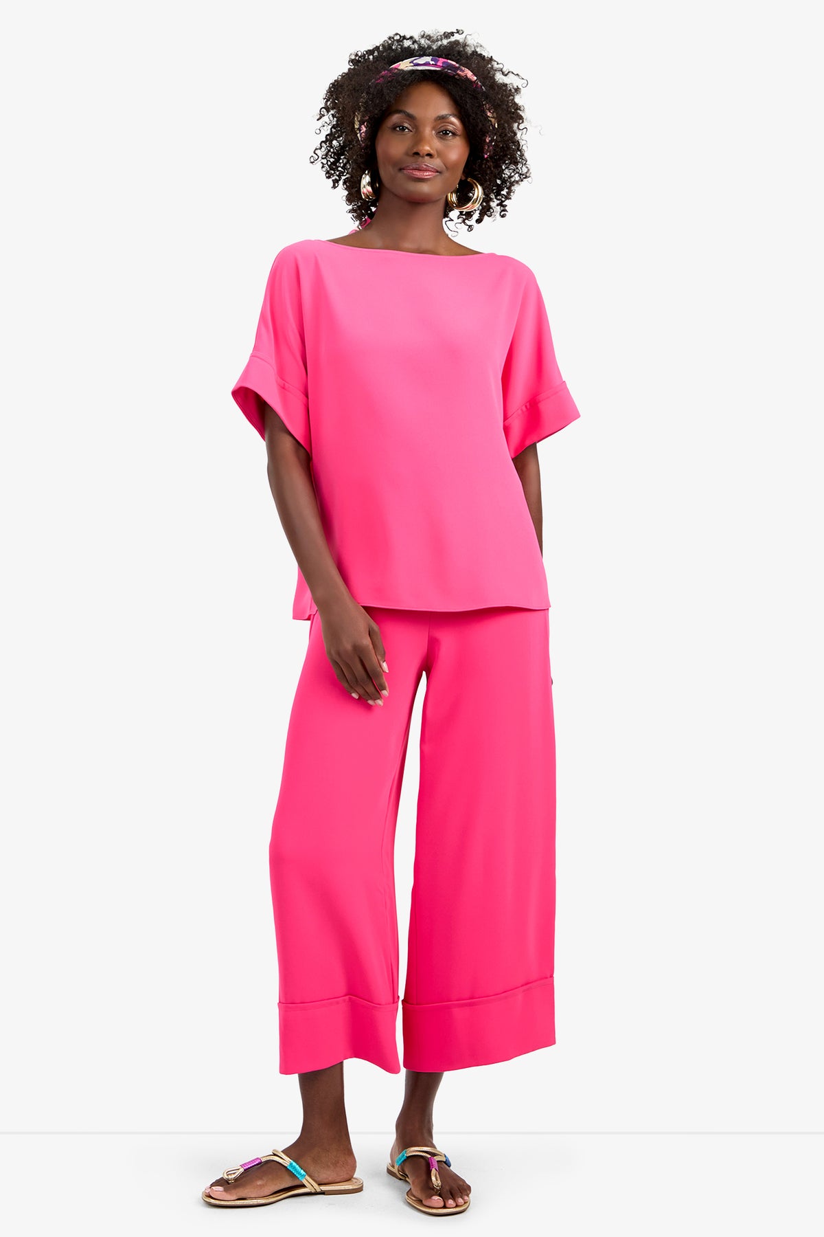 A woman with curly hair and a pink headband smiles, wearing the SOFIE TOP—a loose, bright pink short-sleeve top in soft 100% polyester—paired with matching wide-leg pants and colorful sandals, set against a plain white background.